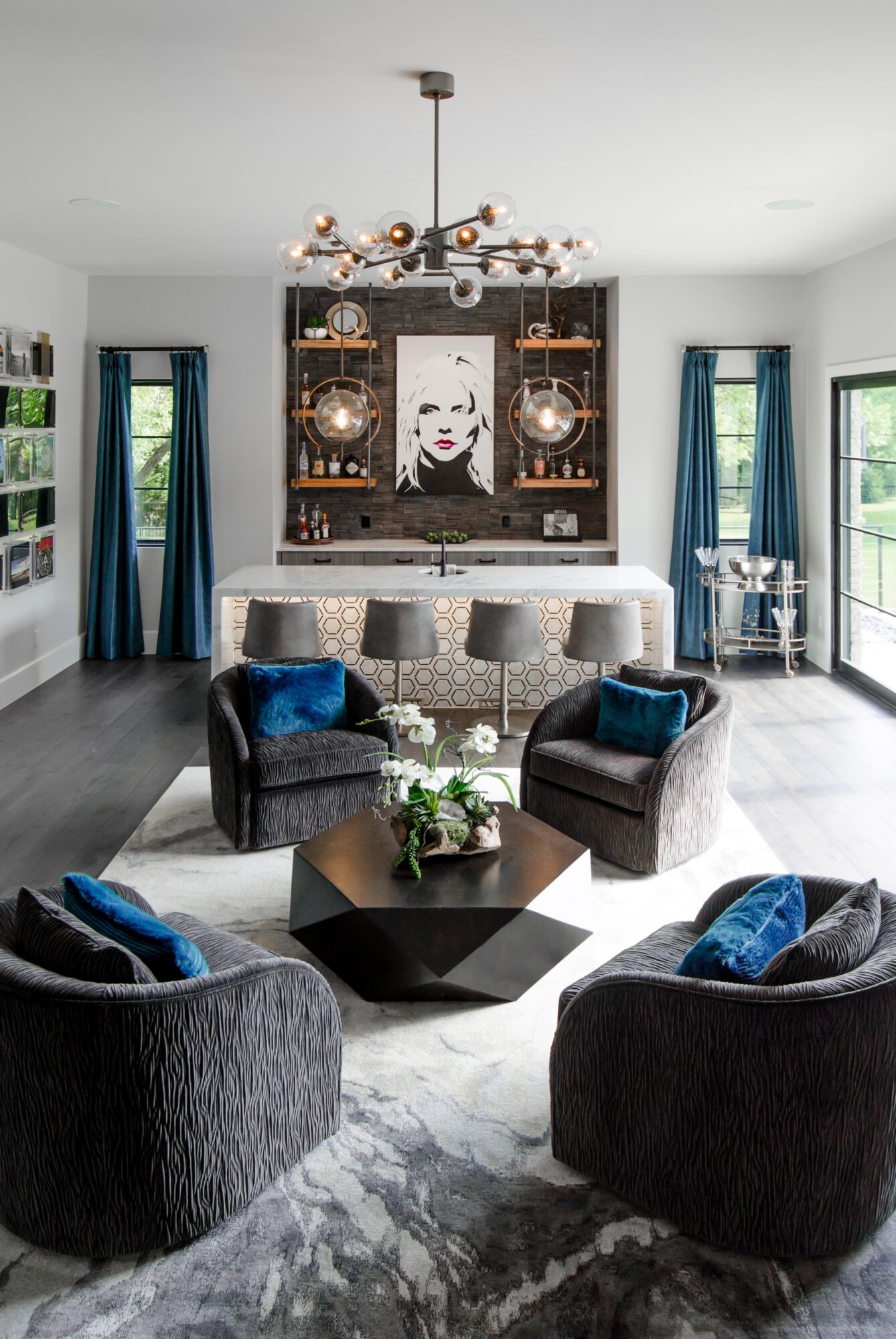 Living room with dark furniture, a chandelier, and large windows with blue curtains, decorated with plants and artwork.