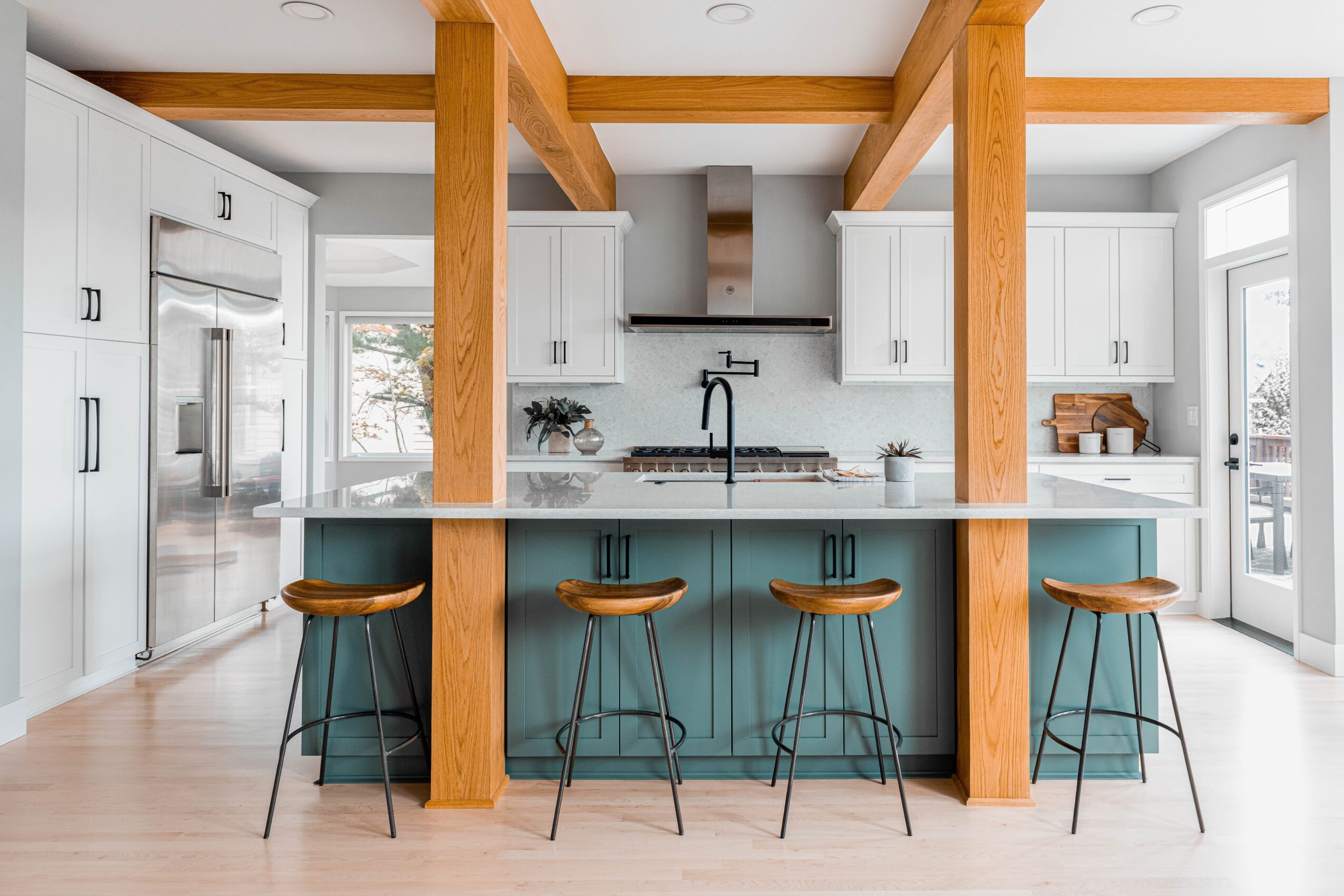 Modern kitchen with white cabinets, a large island with blue base, four bar stools, and wooden beams, bright natural light.
