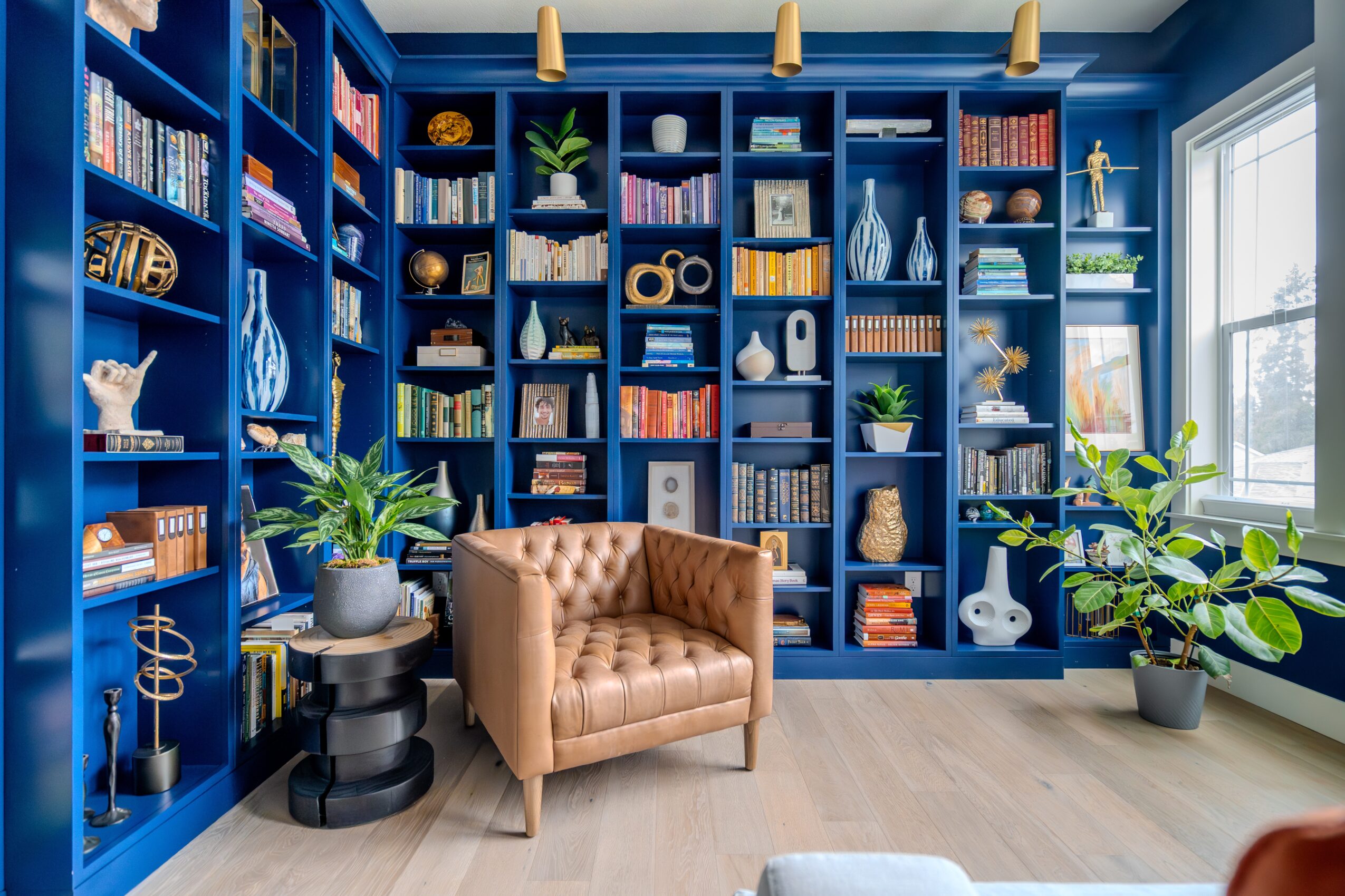 Blue bookshelf filled with books and decorative items, tan armchair, potted plants, window with natural light, light wood floor