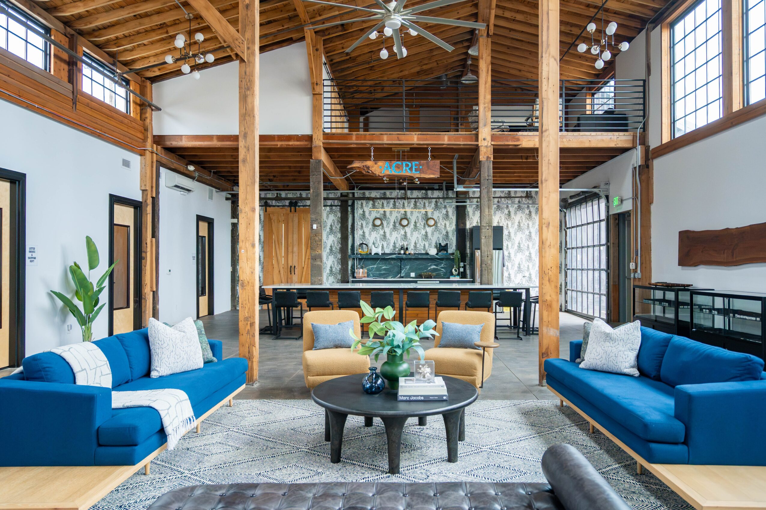 Interior of a spacious, wooden-ceiling lounge with two blue sofas, a round coffee table with flowers, and large windows.
