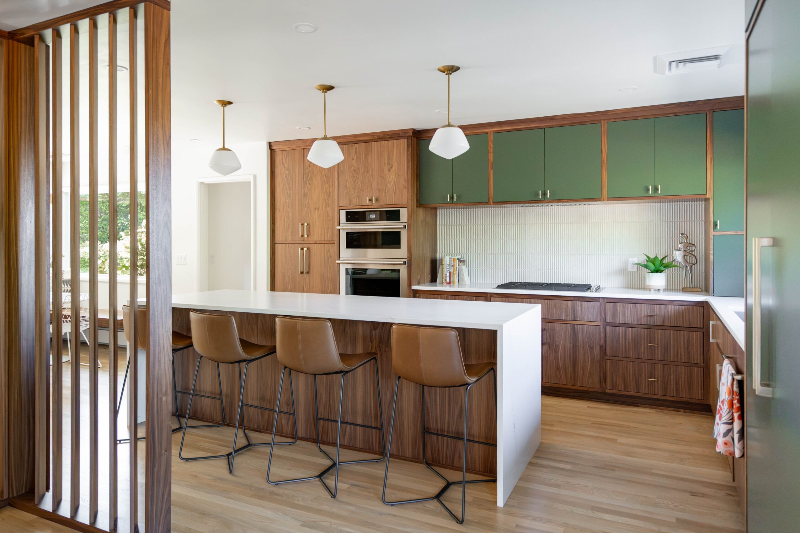 Modern kitchen with green cabinets, wooden accents, and a white island with four chairs, pendant lights, and a window view.