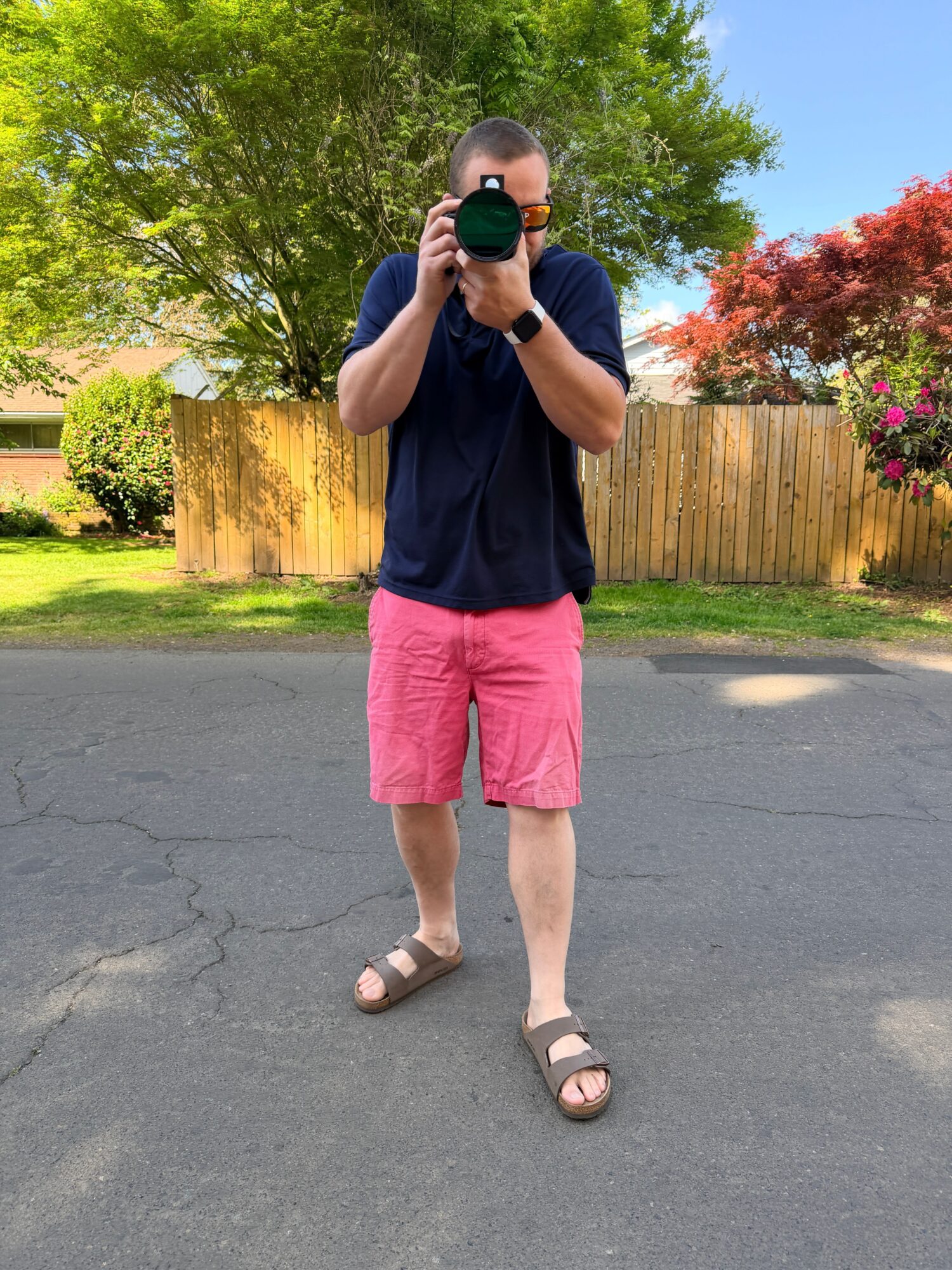 Person taking a selfie outdoors, wearing a navy shirt, pink shorts, and sandals, standing on a paved surface with trees and a wooden fence in background.