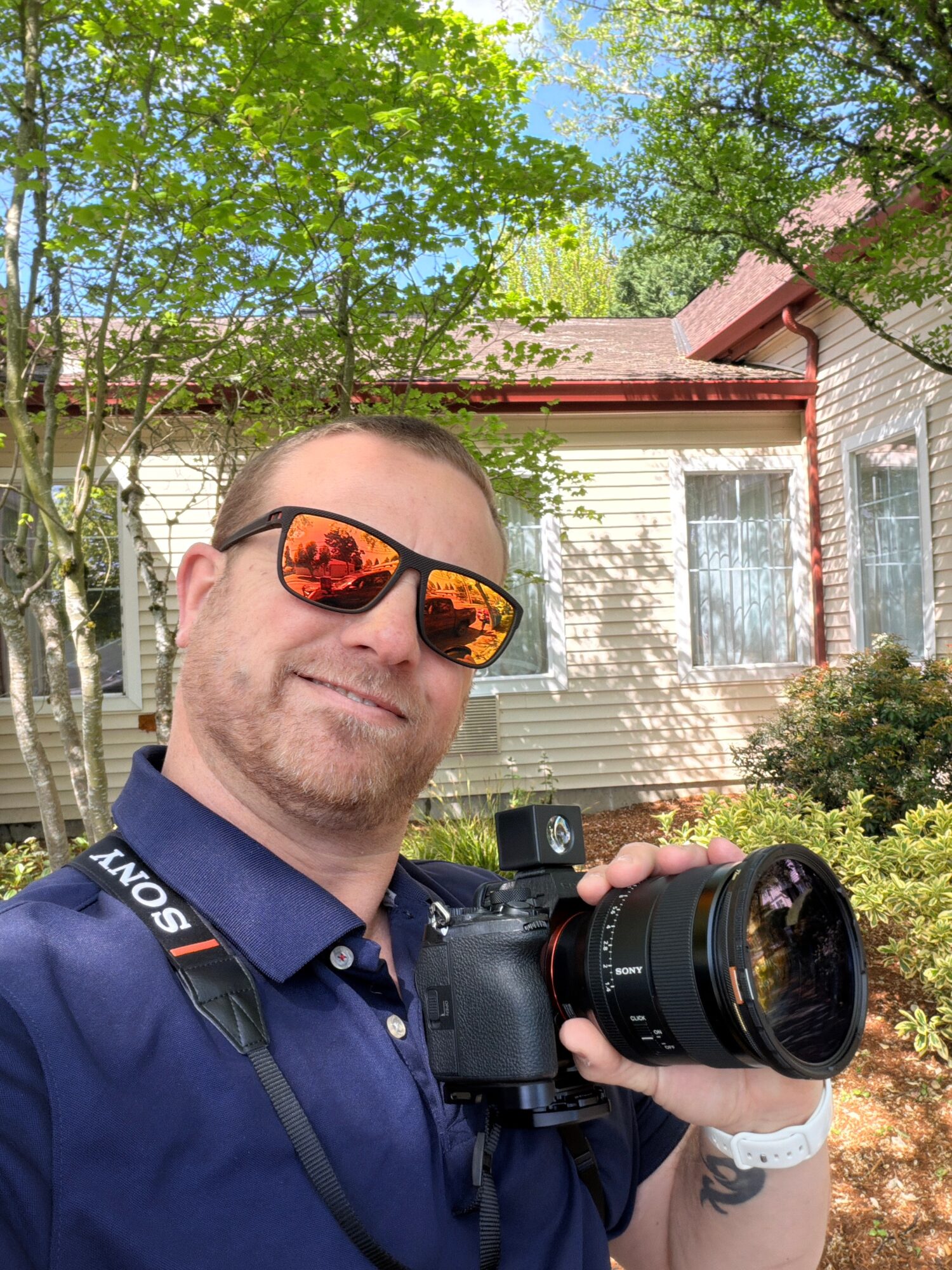 Man wearing sunglasses and a blue shirt holding a camera outdoors in front of a house with trees and bushes