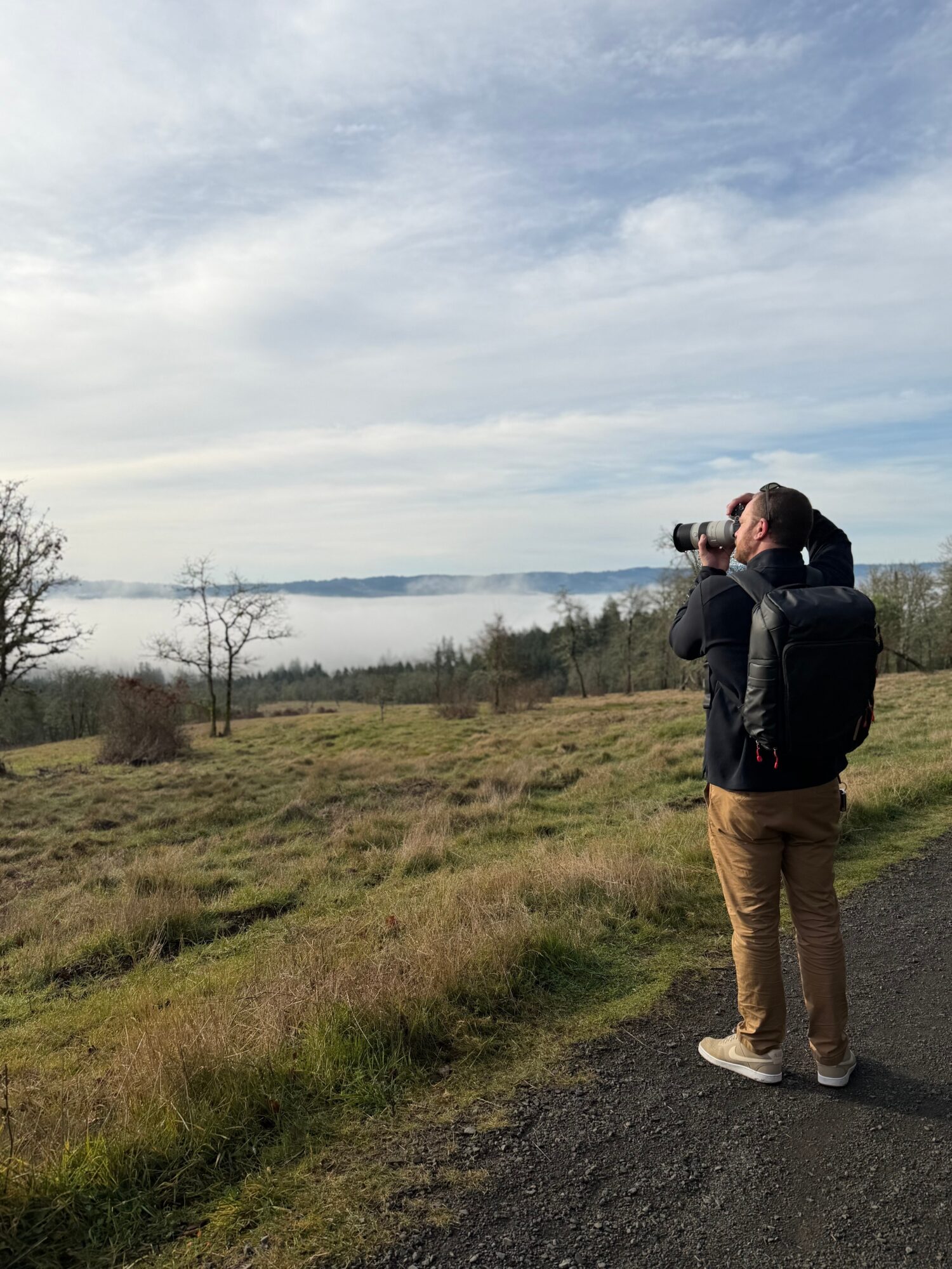Person standing on a path, looking at a landscape with water and trees, holding a camera or binoculars, cloudy sky