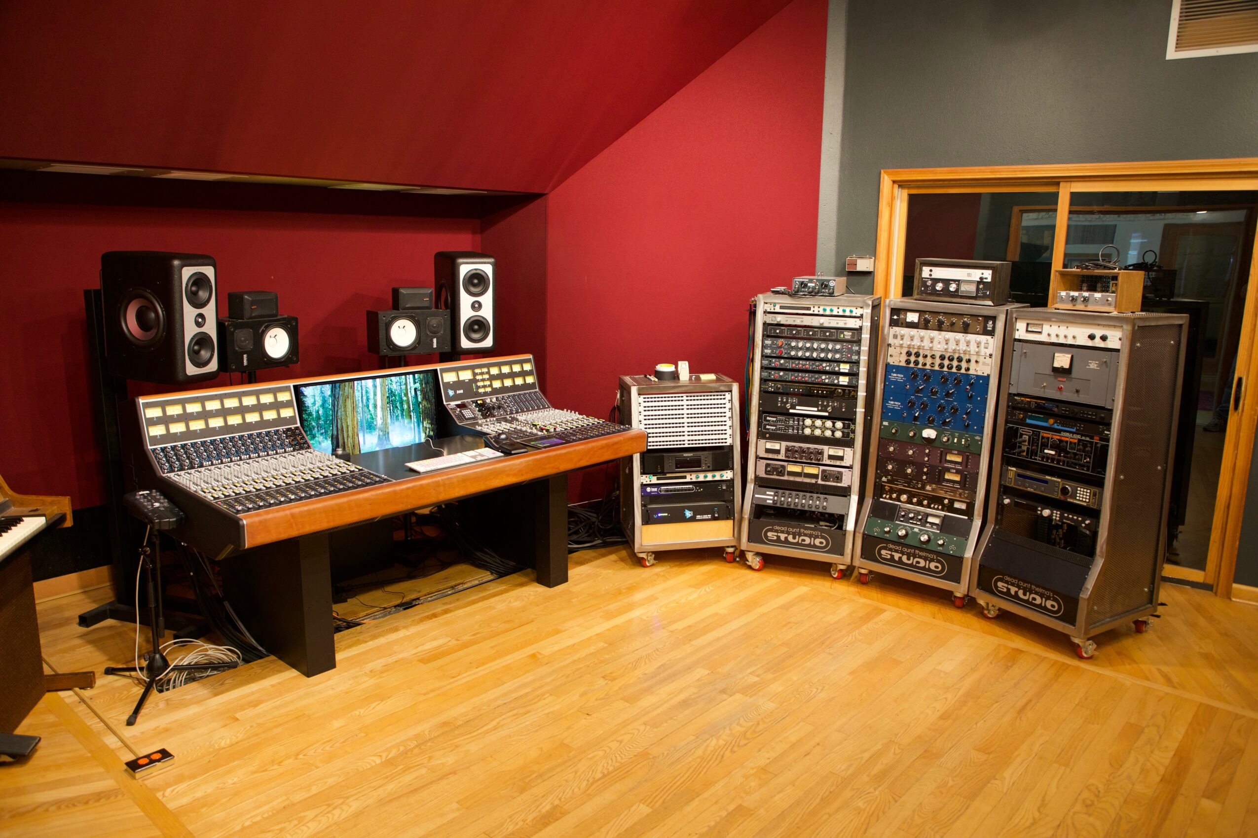 Recording studio with mixing console, speakers, and audio equipment on racks, in a room with red and gray walls and wooden floor.