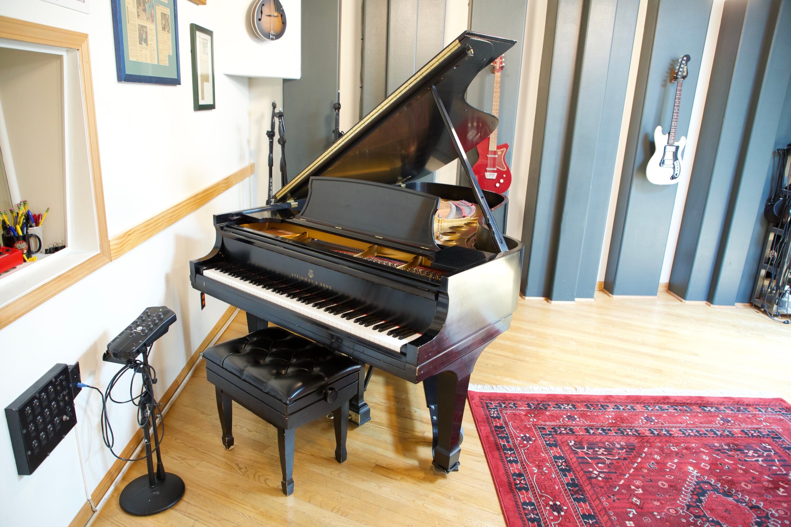 Black grand piano with open lid in a music room, with a stool, guitar wall hangings, and a red rug.