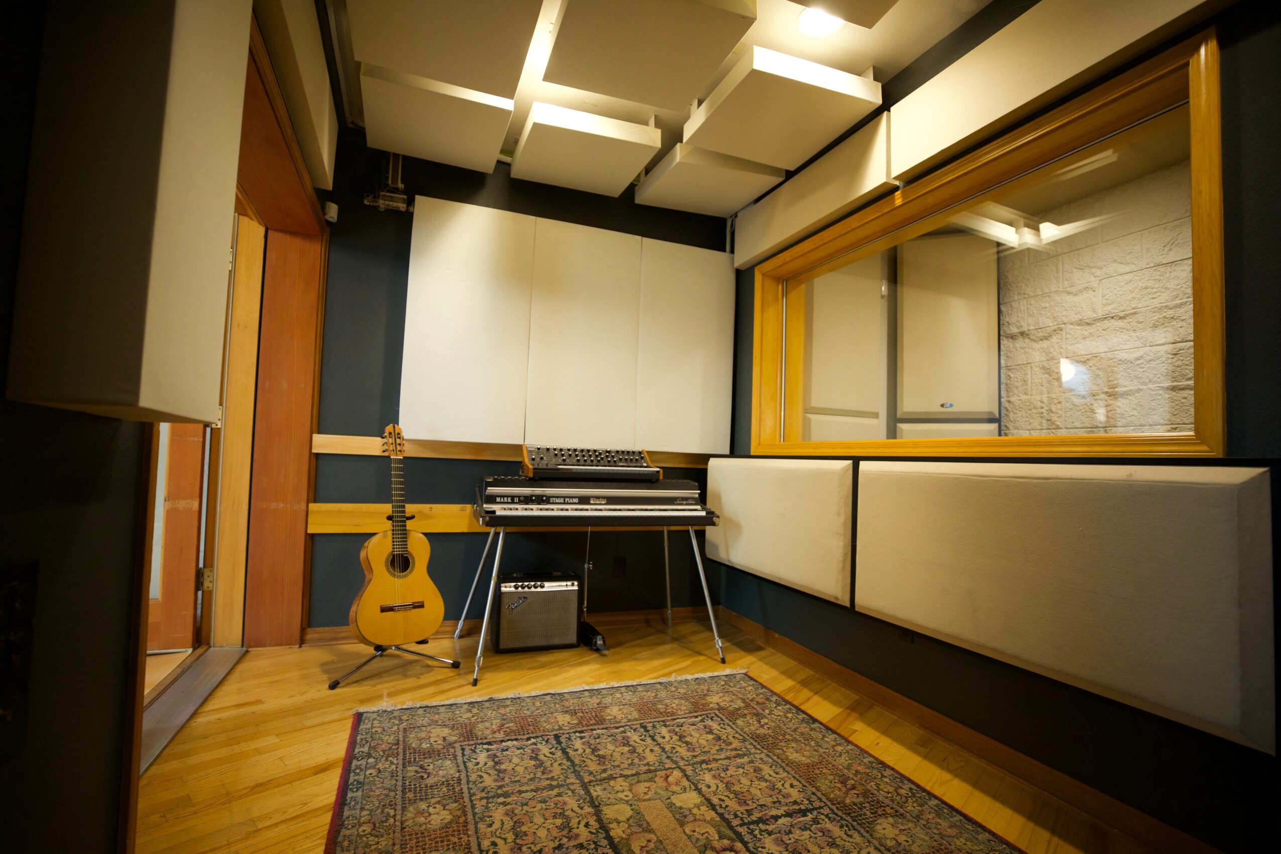 Recording studio with a guitar, keyboard, and sound equipment, viewed through a window, with a rug on wooden floor.
