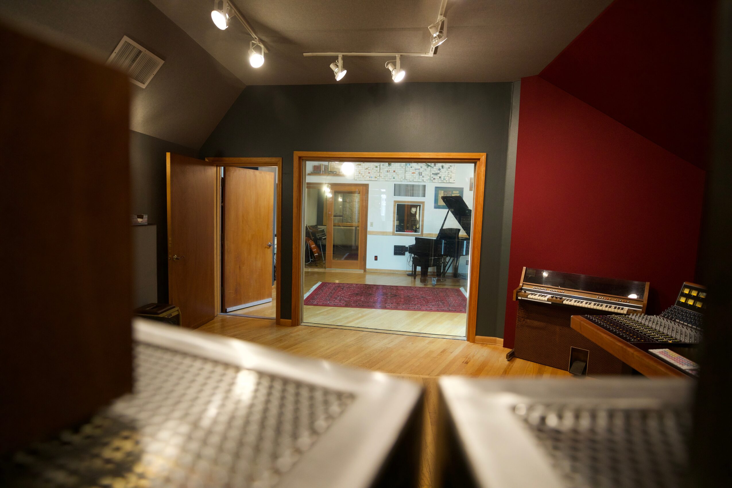 Recording studio with a window, piano, and wooden furniture, illuminated by ceiling lights, with a red and gray wall.