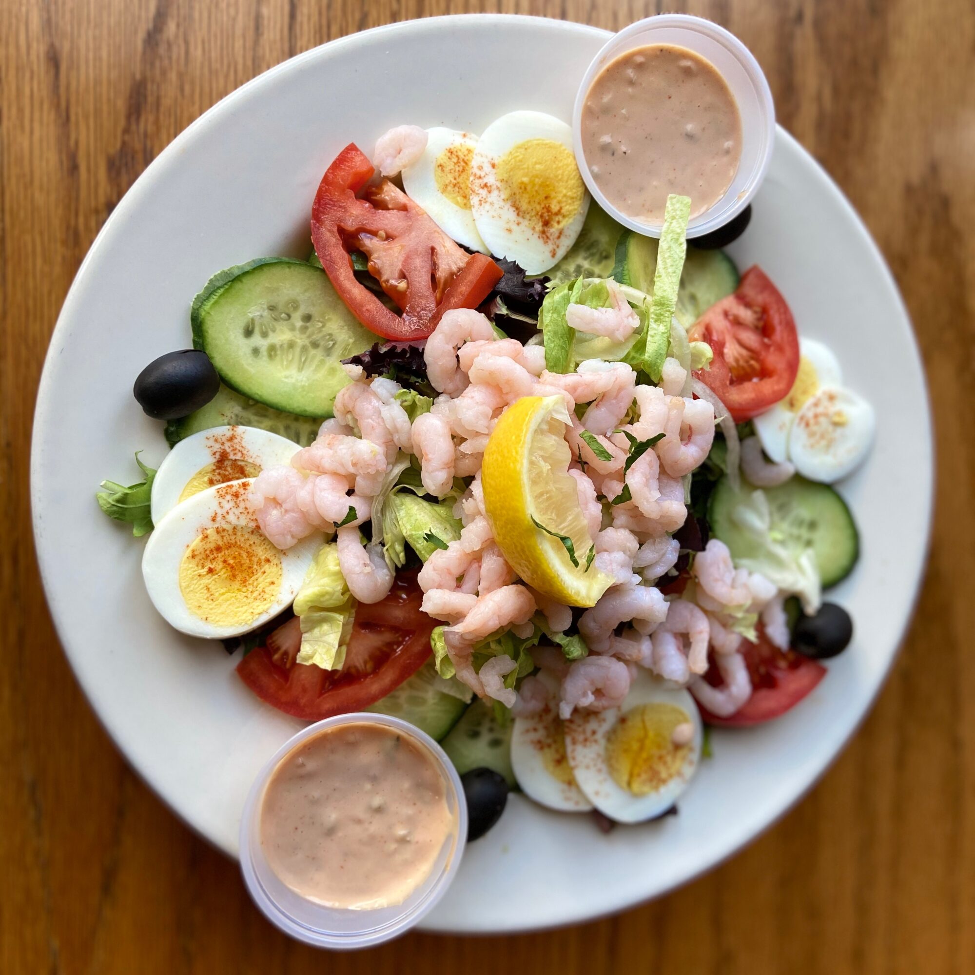 Salad with shrimp, cucumber, tomato, boiled eggs, lemon, olives, and dressing on a white plate.