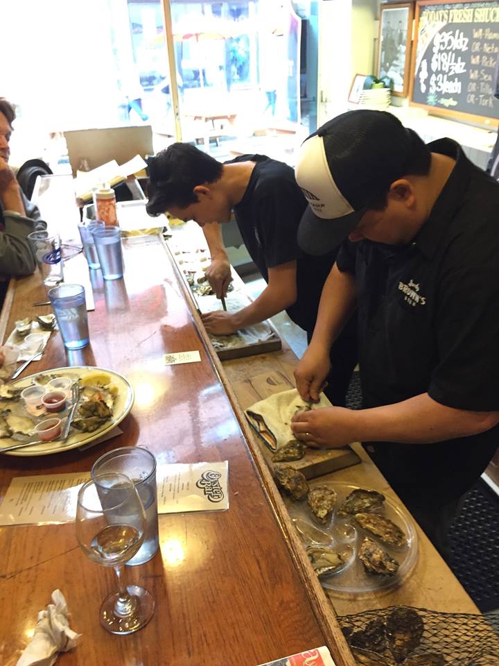 People standing at a wooden counter preparing food, with plates and glasses on the table, in a bright restaurant interior.