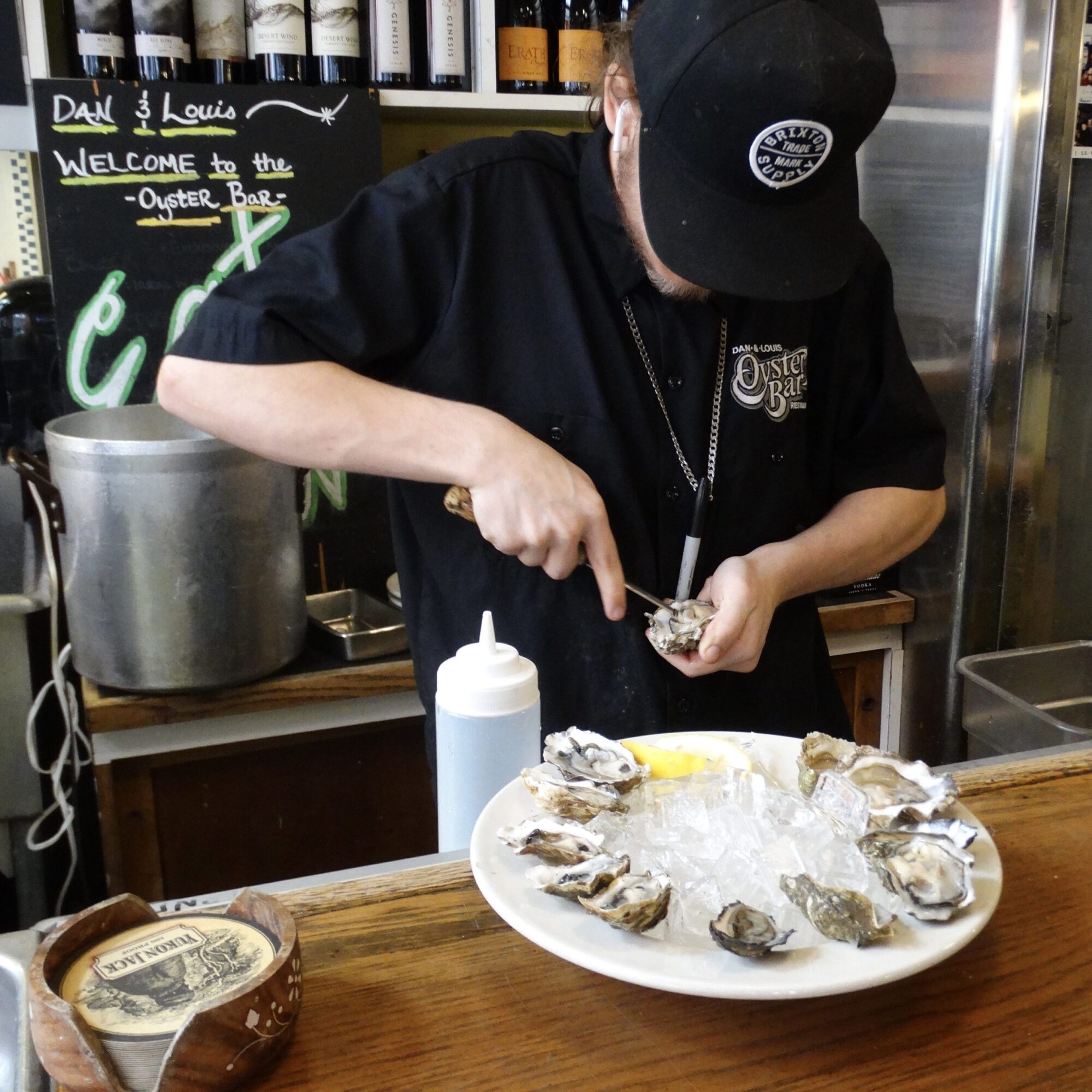 Person shucking oysters at a bar, with a plate of oysters on ice in front. Person wears a black cap and shirt.