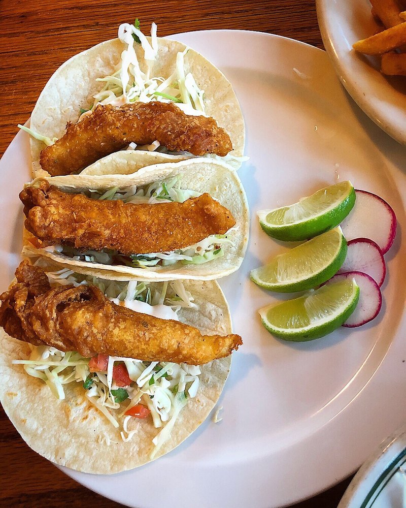 Three fish tacos with shredded lettuce and fried fish in soft tortillas, served with lime and onion slices on a white plate.