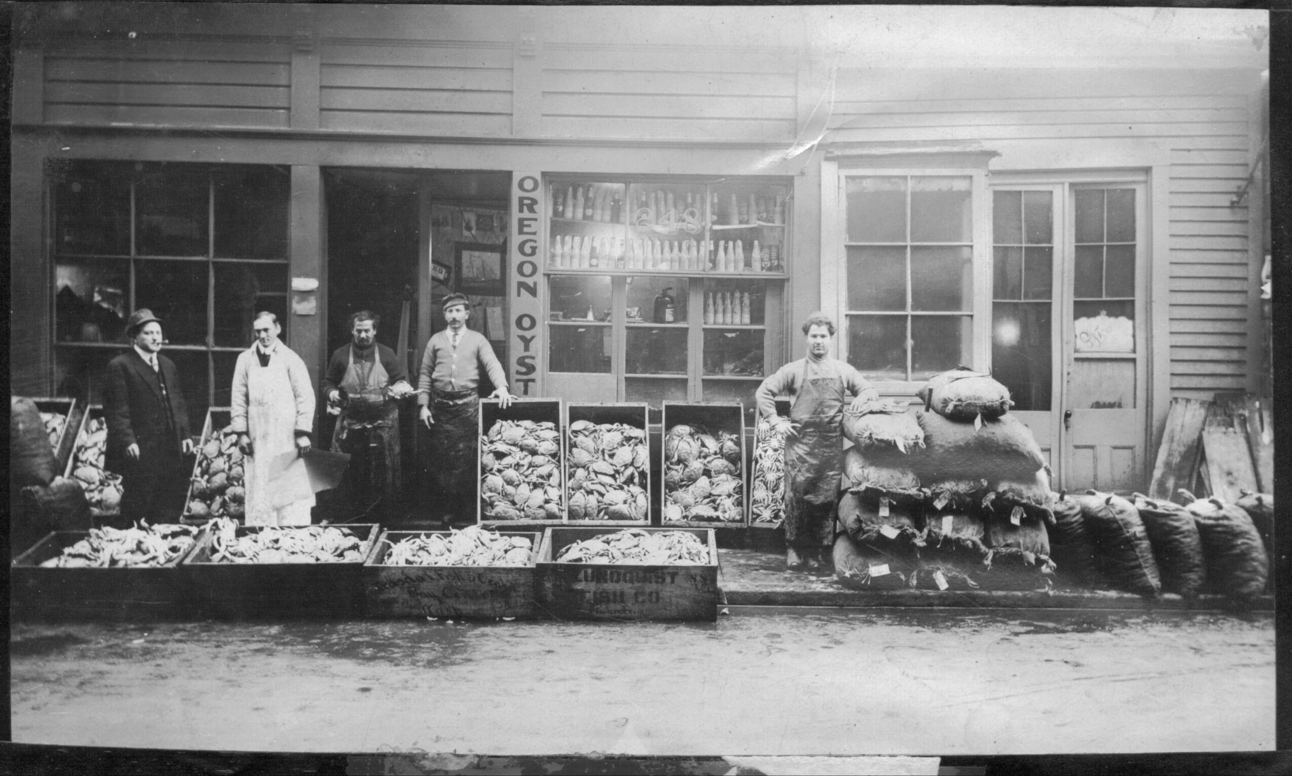 Black and white photo of people standing outside a store with large windows, selling produce and goods, in front of a building.