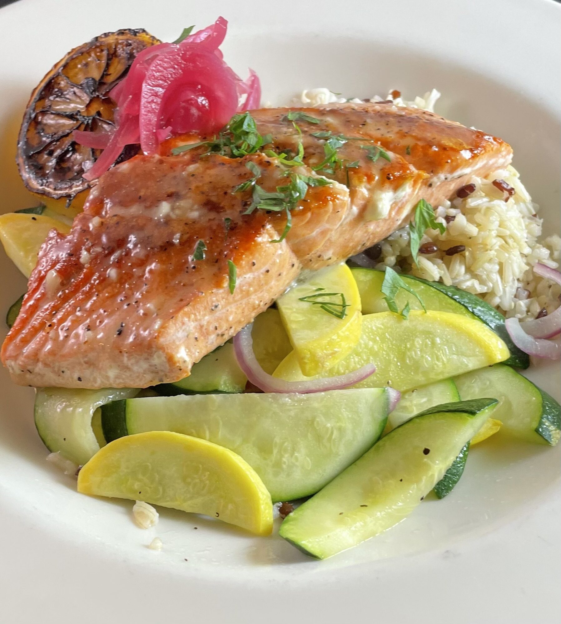 Cooked salmon fillet on a bed of rice with cucumber, yellow squash, and pickled vegetables, garnished with herbs.
