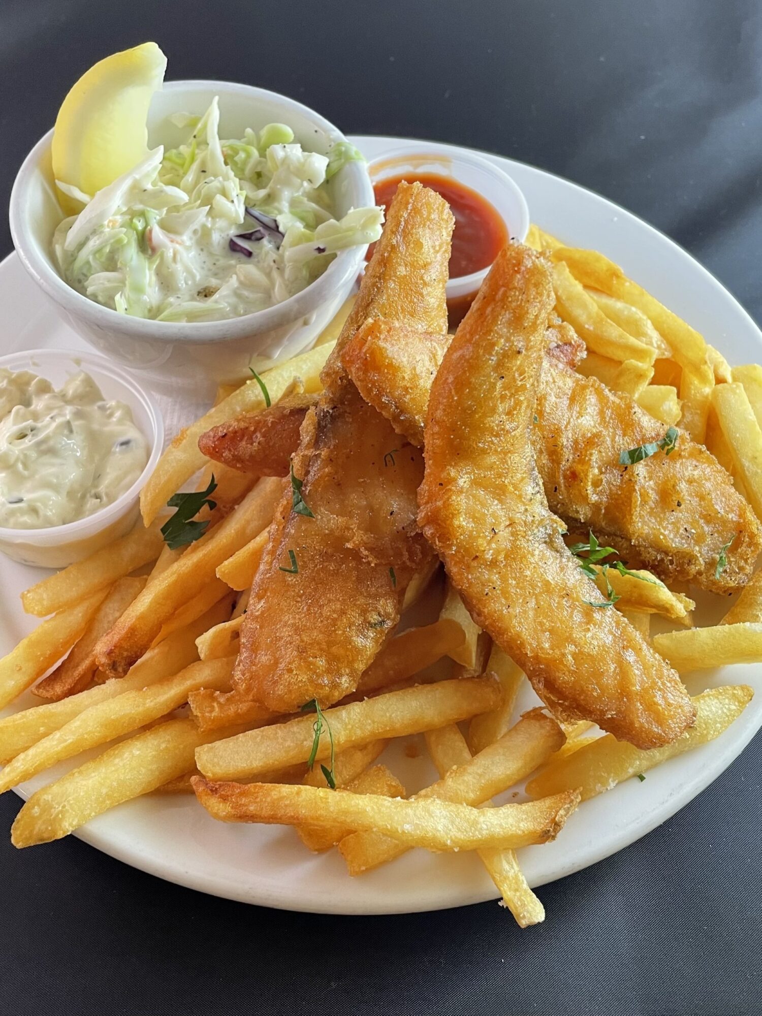 Plate with fried fish, French fries, salad, lemon wedge, and tartar sauce, on a dark surface.