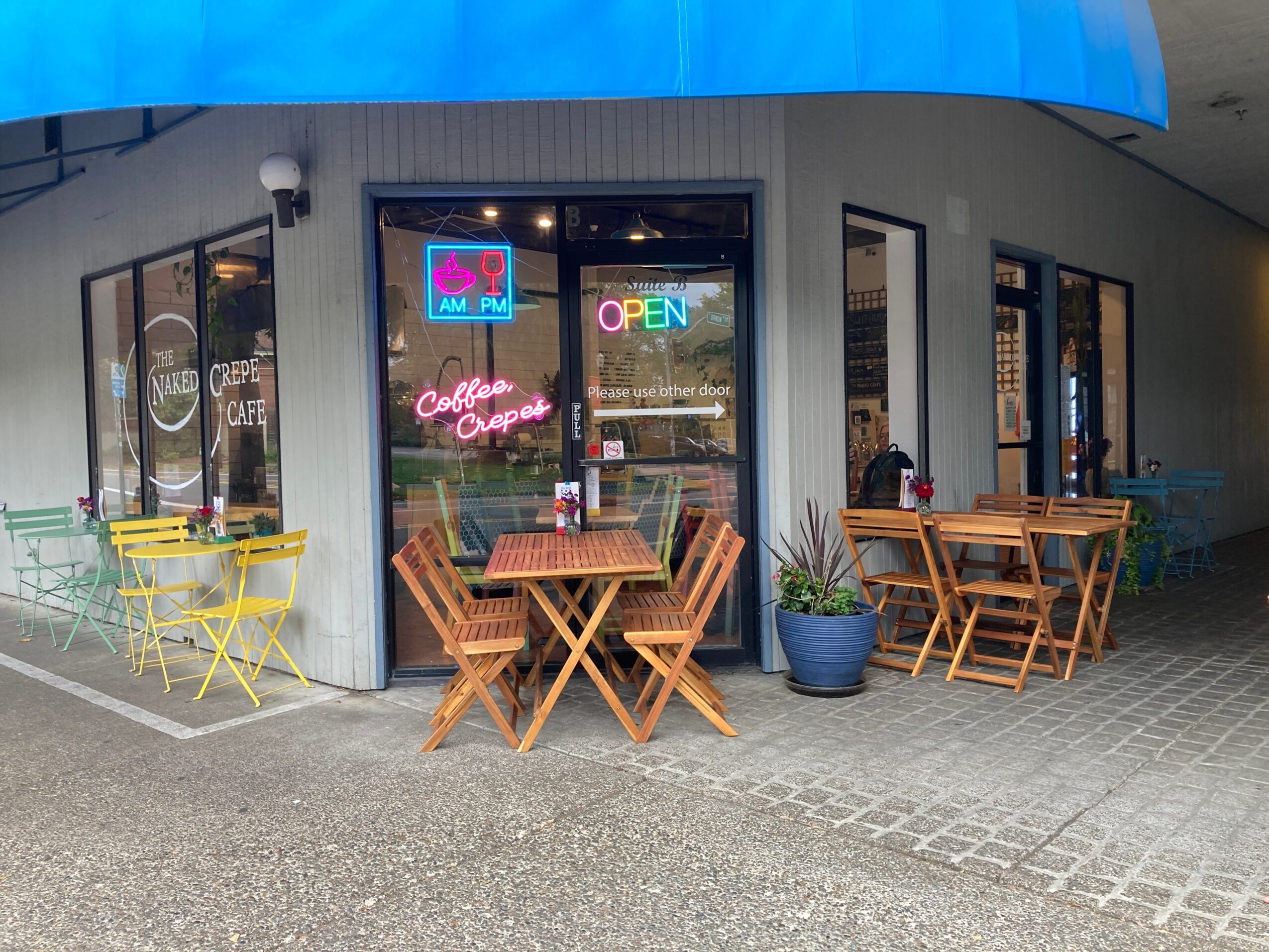 Cafe storefront with outdoor tables and chairs, neon signs, and a potted plant outside.
