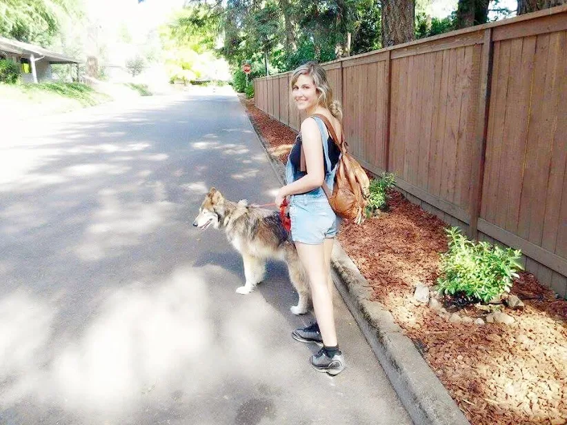 Girl with a backpack standing on a sidewalk next to a dog, with a wooden fence and trees in the background.