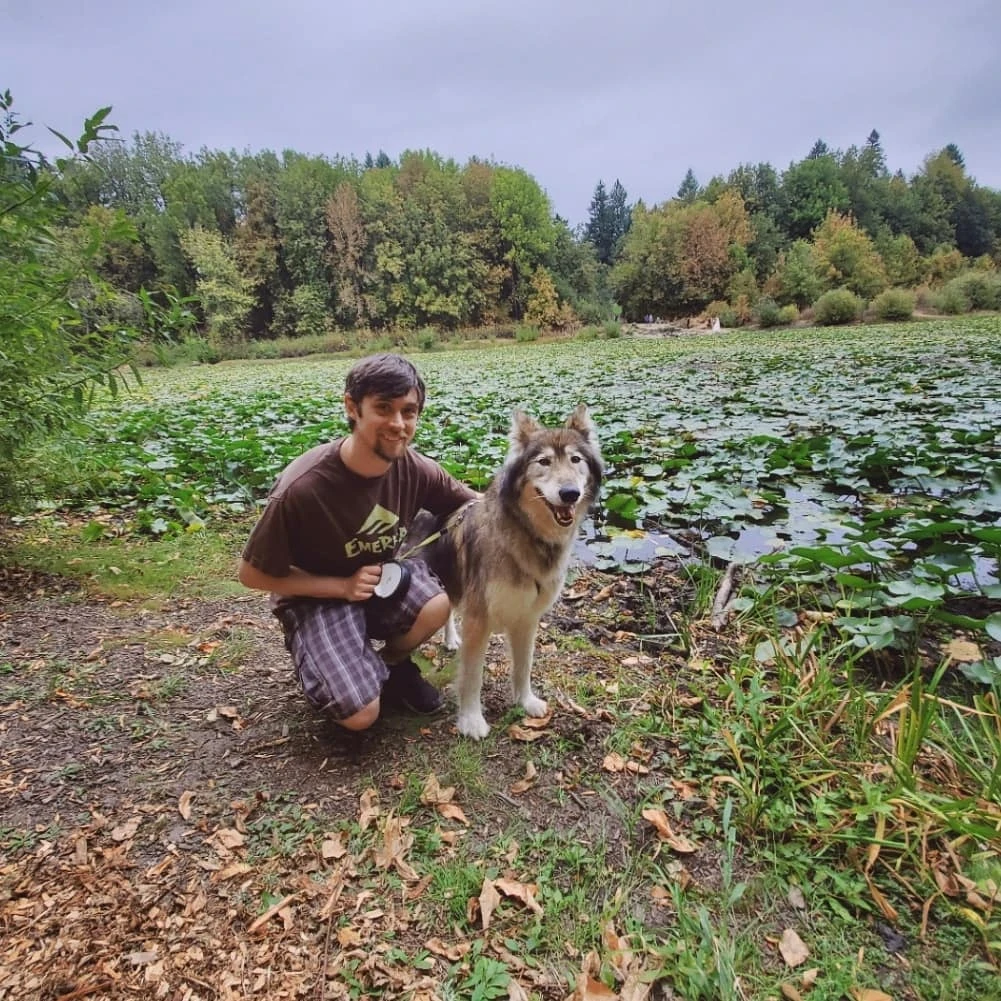 A young man crouching beside a large dog near a pond with lily pads, trees in the background, overcast sky.
