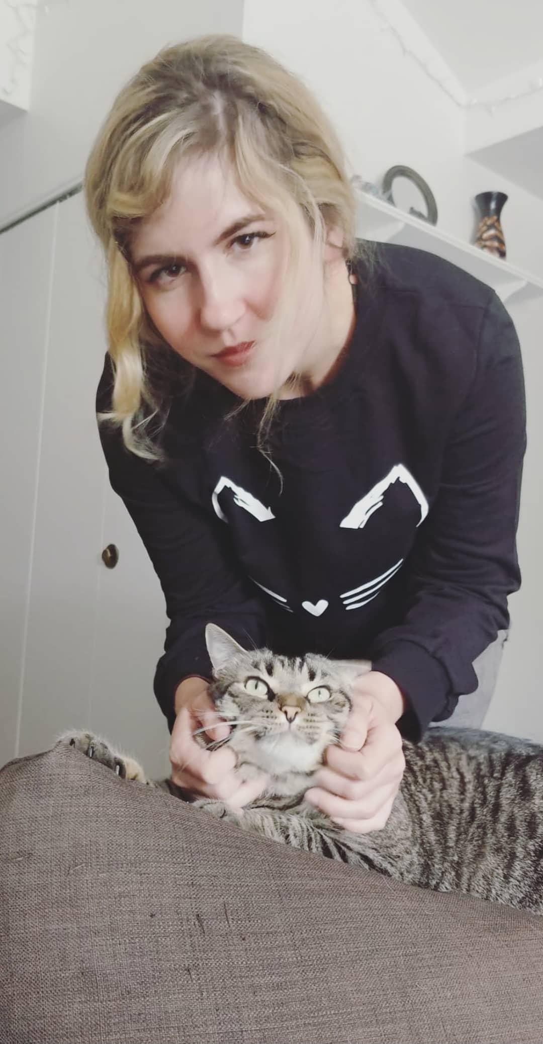 Person with blonde hair holding a small tabby cat, both looking at the camera, in a room with white cabinets
