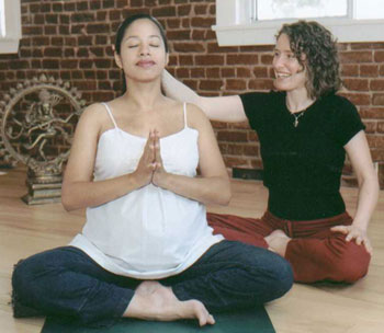 Two women practicing yoga indoors, one meditating with hands in prayer position, the other smiling and touching her shoulder.
