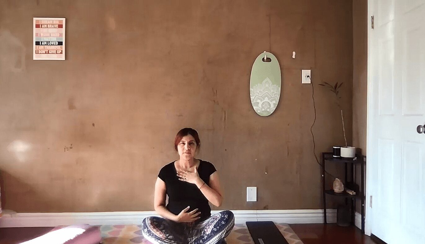 Person sitting cross-legged on a yoga mat in a room with a brown wall, mirror, and small shelf, holding their stomach.