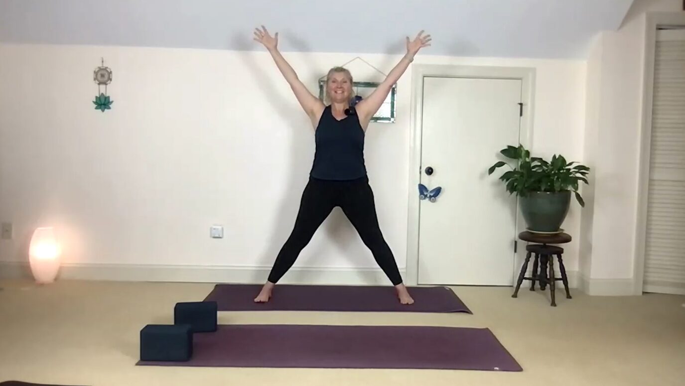 Person standing on yoga mat with arms raised in a spacious room, next to a potted plant and a door, with a small lamp on the floor.