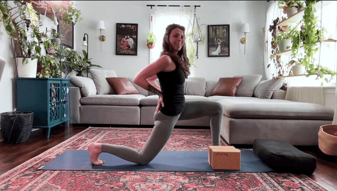 Woman practicing yoga on a mat in a living room with a sofa, plants, and framed pictures, kneeling with hands on hips.