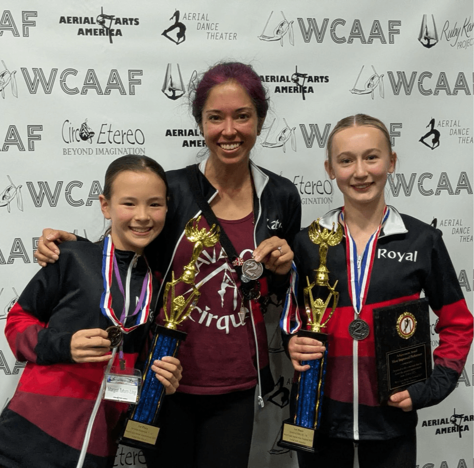 Three smiling women holding trophies and medals, standing in front of a backdrop with logos and text.