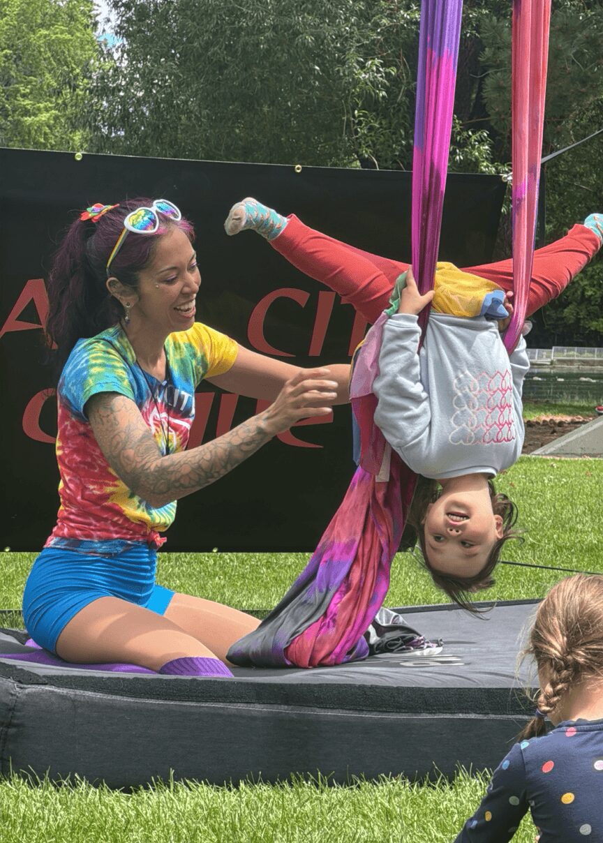 A woman with colorful tie-dye shirt and shorts holds a girl upside down on aerial silks outdoors, with a black banner and trees in background.
