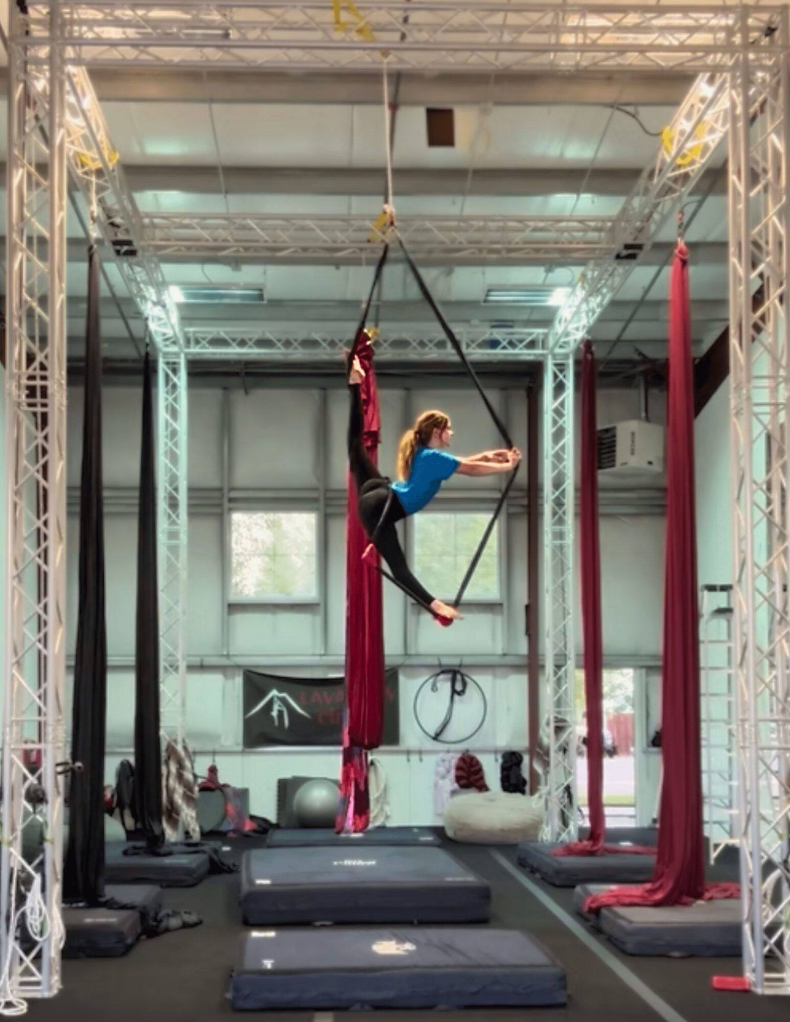 Person performing aerial silk routine on a trapeze in a gym with equipment and mats, high ceiling, and large windows.