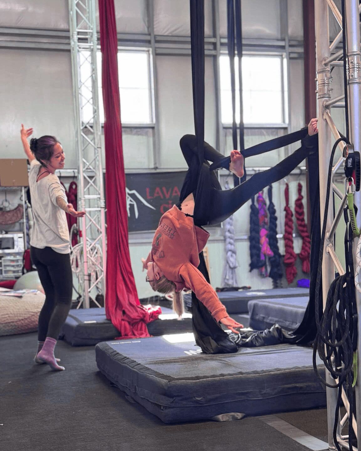 Person hanging upside down from aerial silks, another person standing nearby in a gym with equipment and mats.