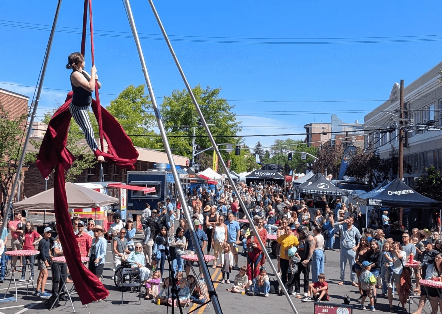 Person on aerial silk performance at outdoor event with crowd and tents, blue sky, and buildings in background.