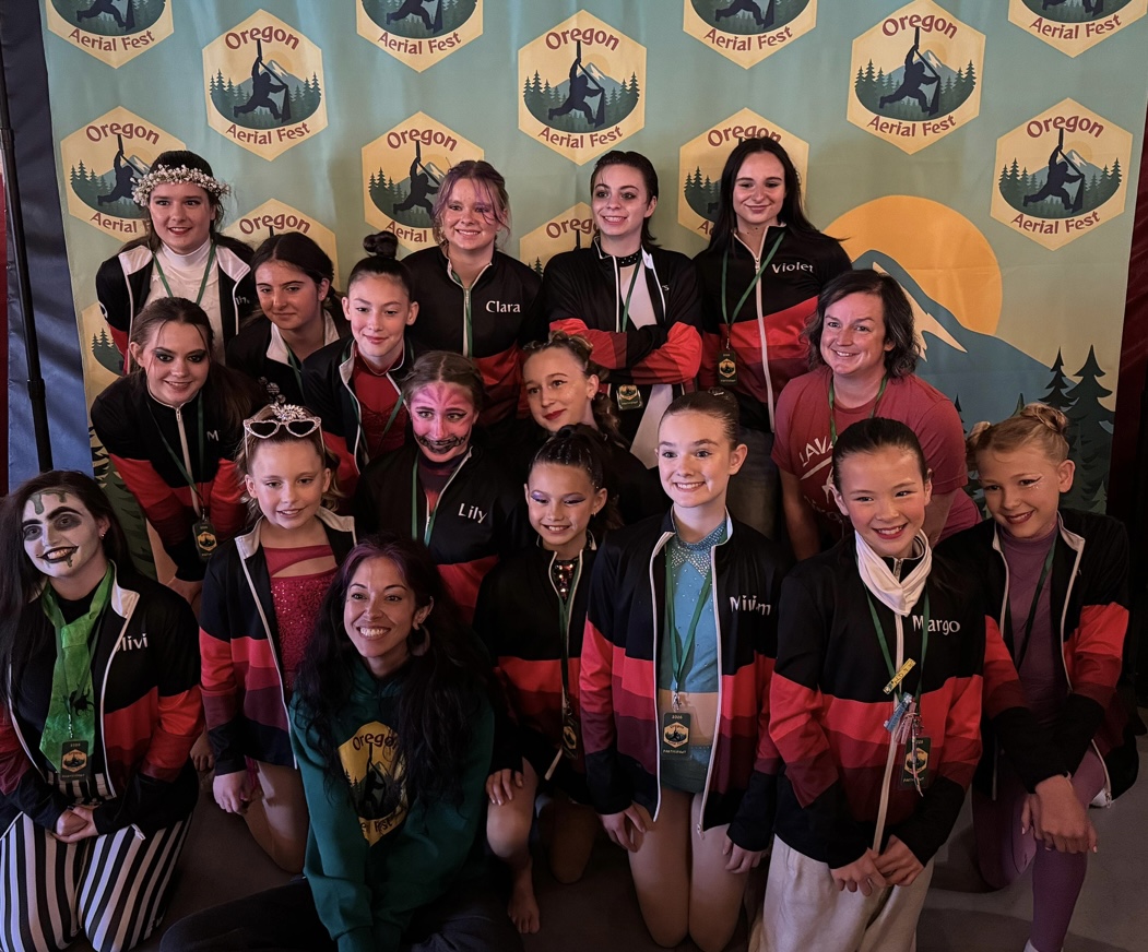 Group of young girls and women posing in front of Oregon-themed backdrop with logos, smiling and wearing medals.