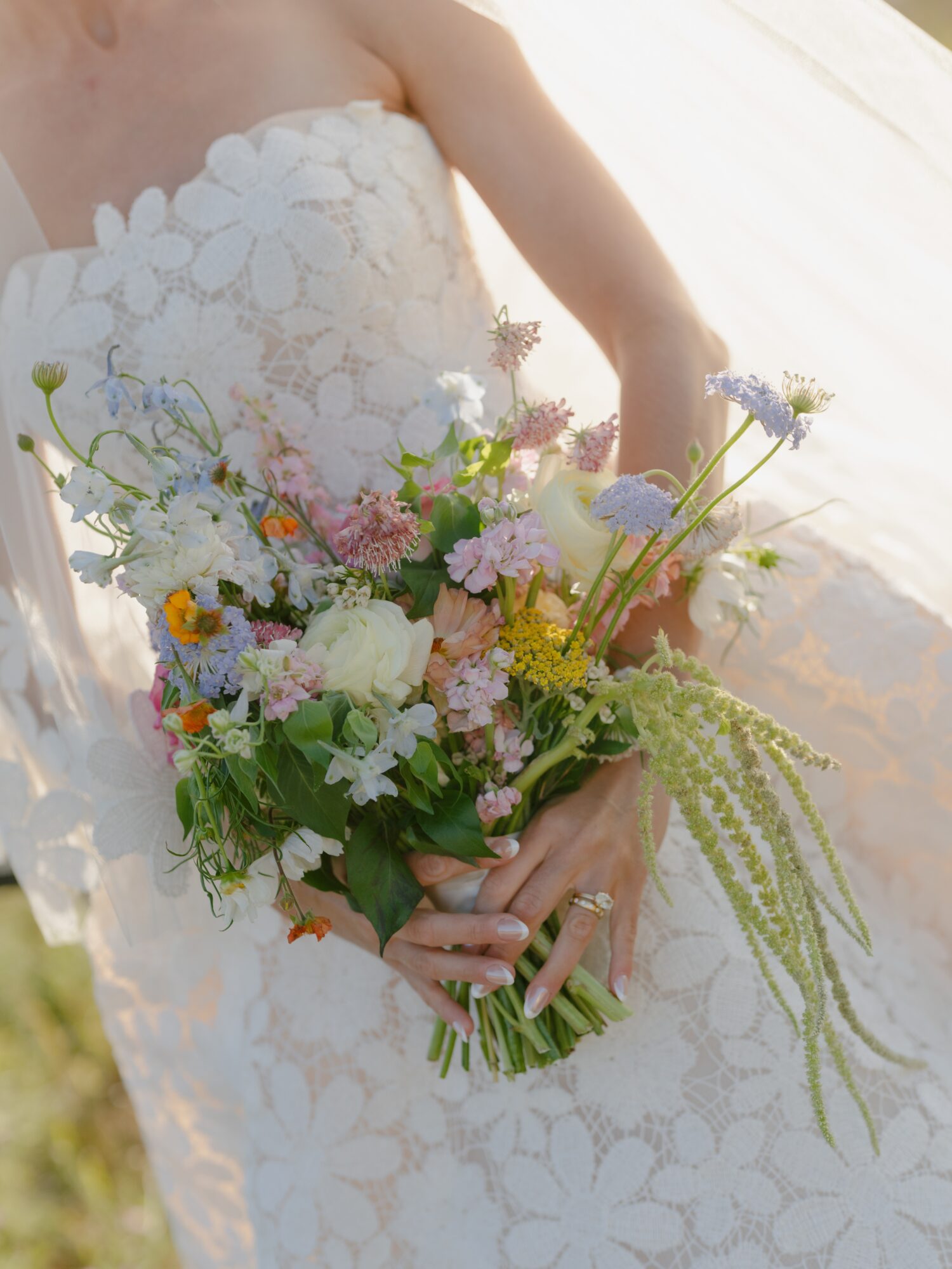 Person in a white lace dress holding a colorful bouquet of flowers, outdoors with sunlight.