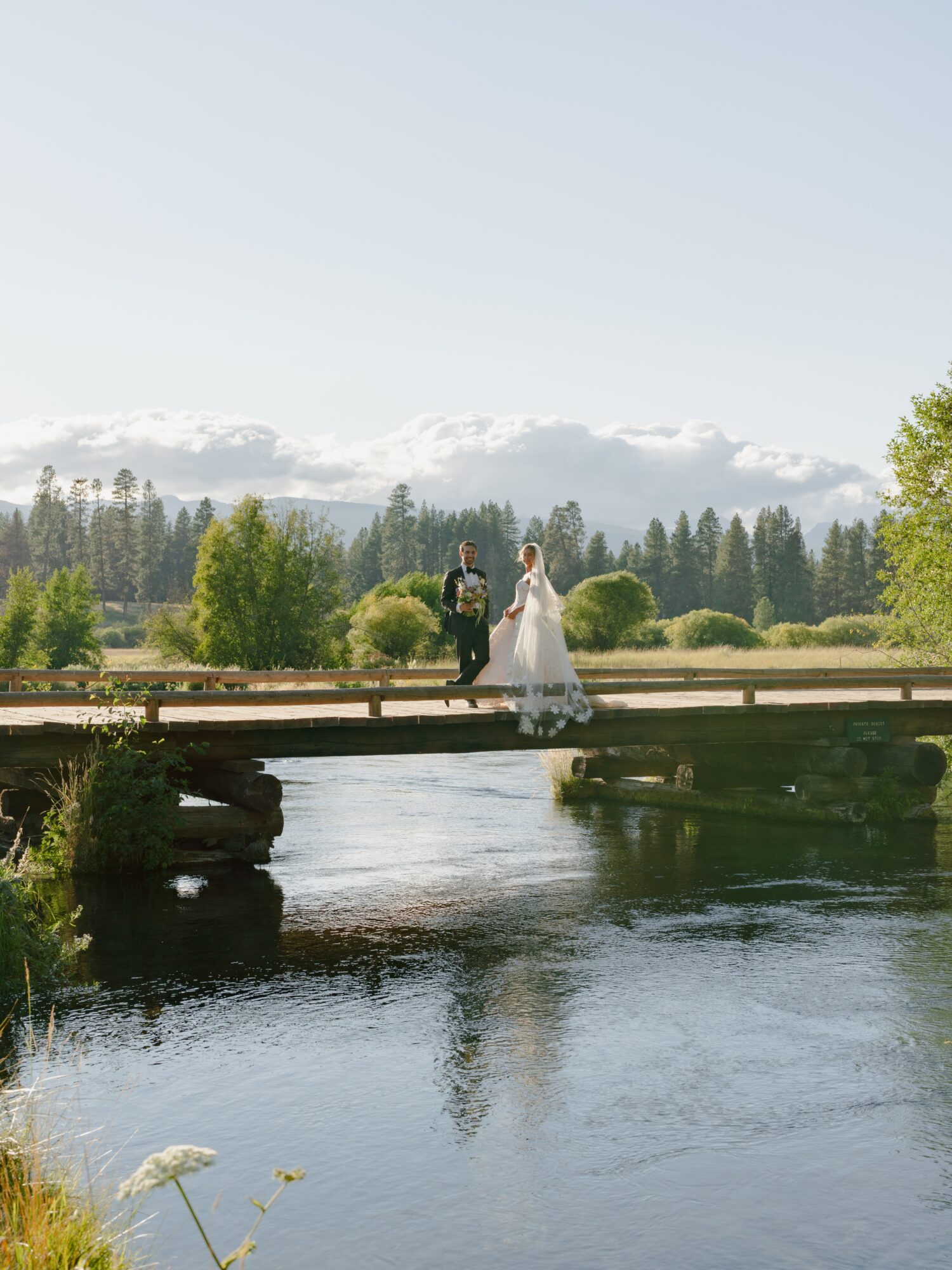 A bride and groom stand on a wooden bridge over a river, surrounded by trees and mountains, under a clear sky.