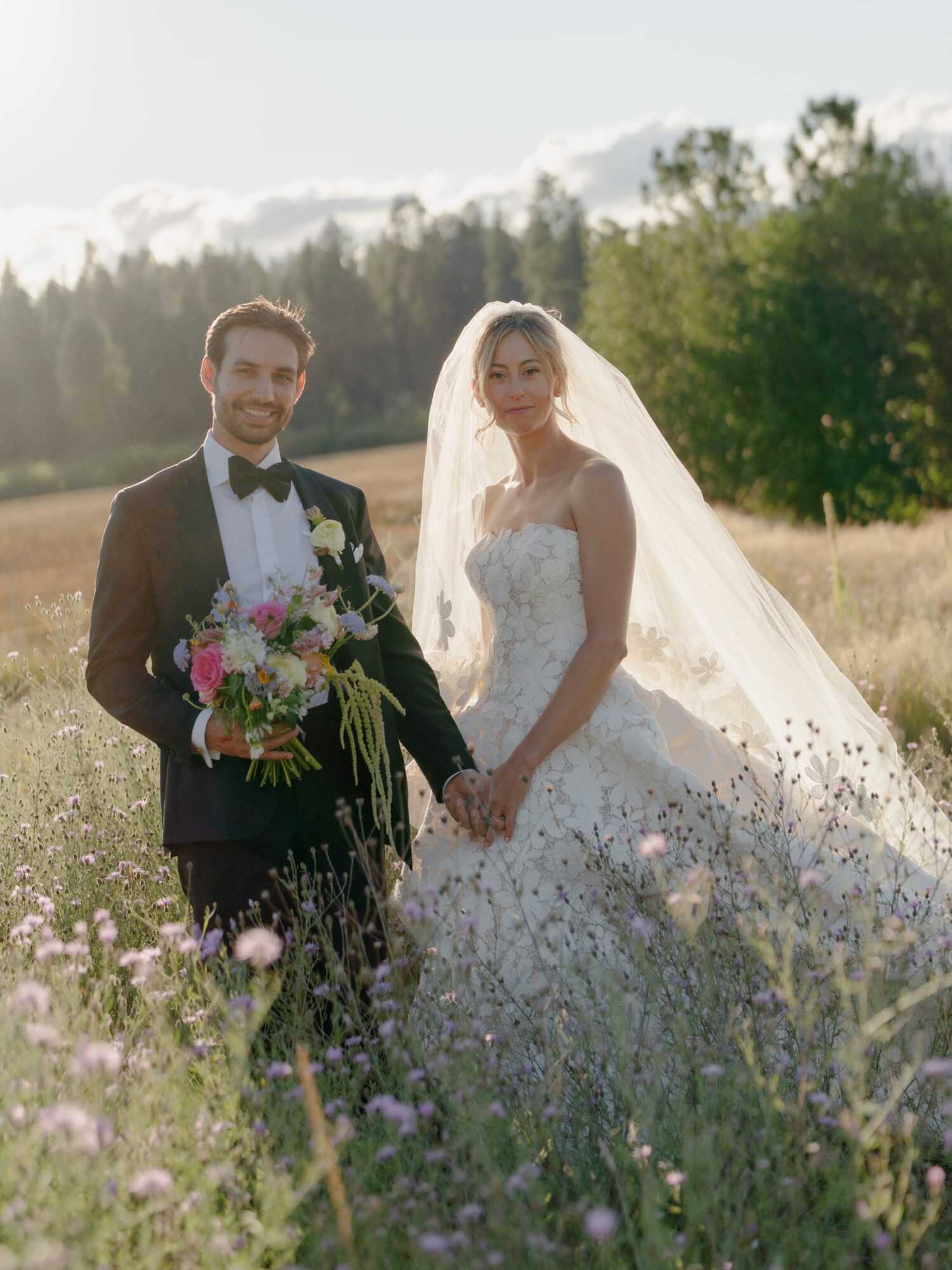 A bride and groom holding hands in a field with trees and sky in the background, sunlight shining on them.