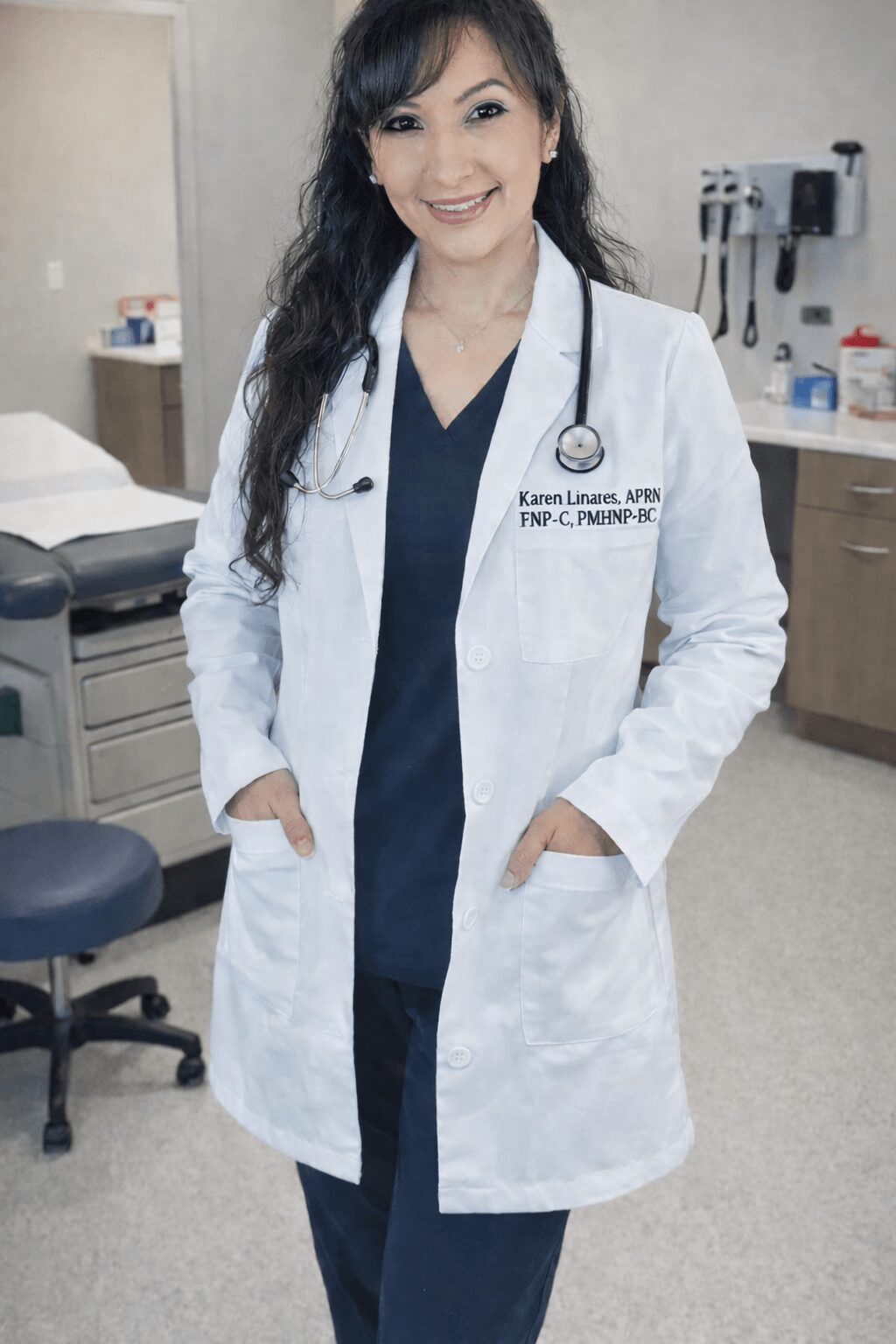 Female doctor in white coat standing in medical office, smiling, hands in coat pockets, stethoscope around neck.