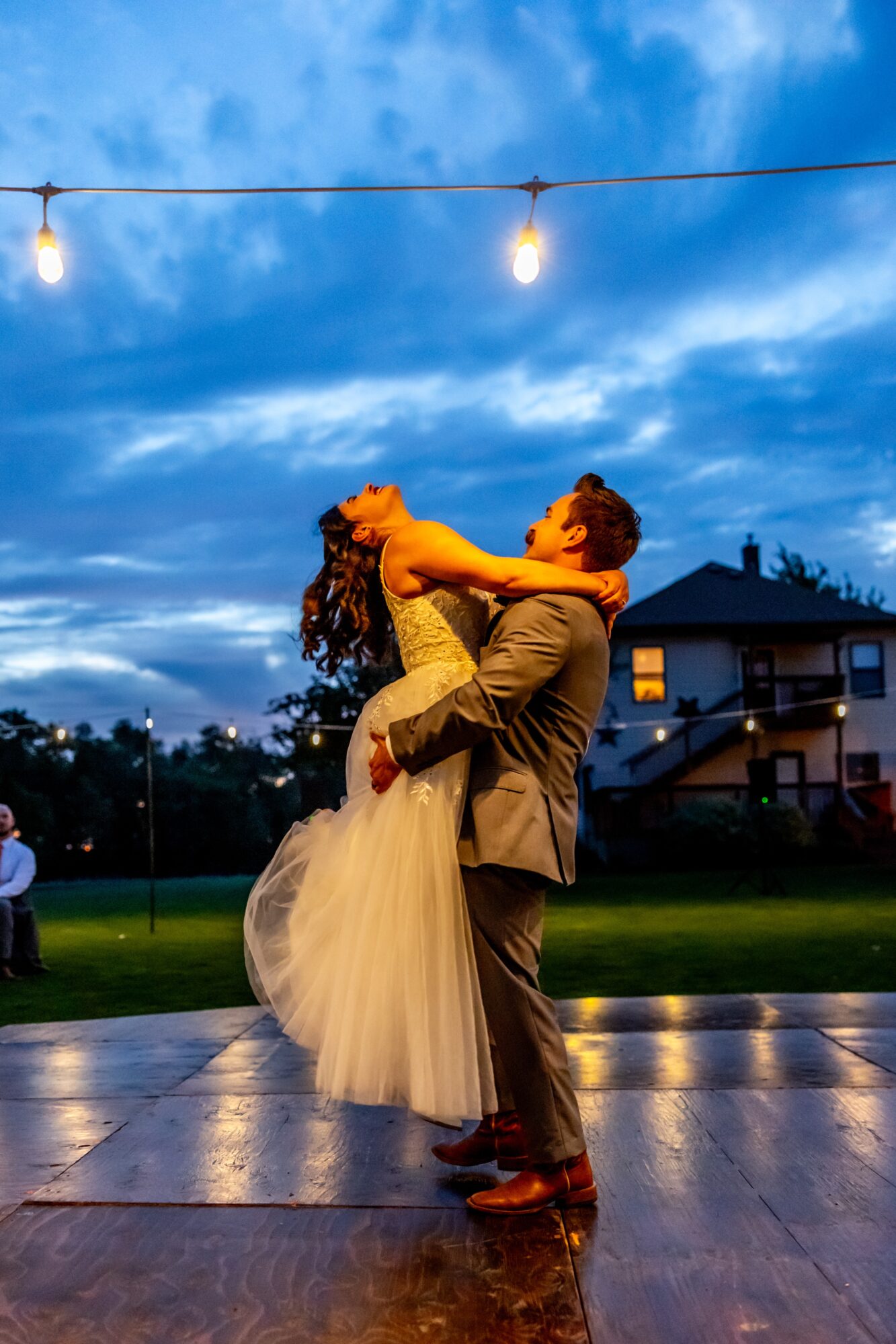 A couple dancing outdoors at dusk, with string lights overhead and a house in the background.
