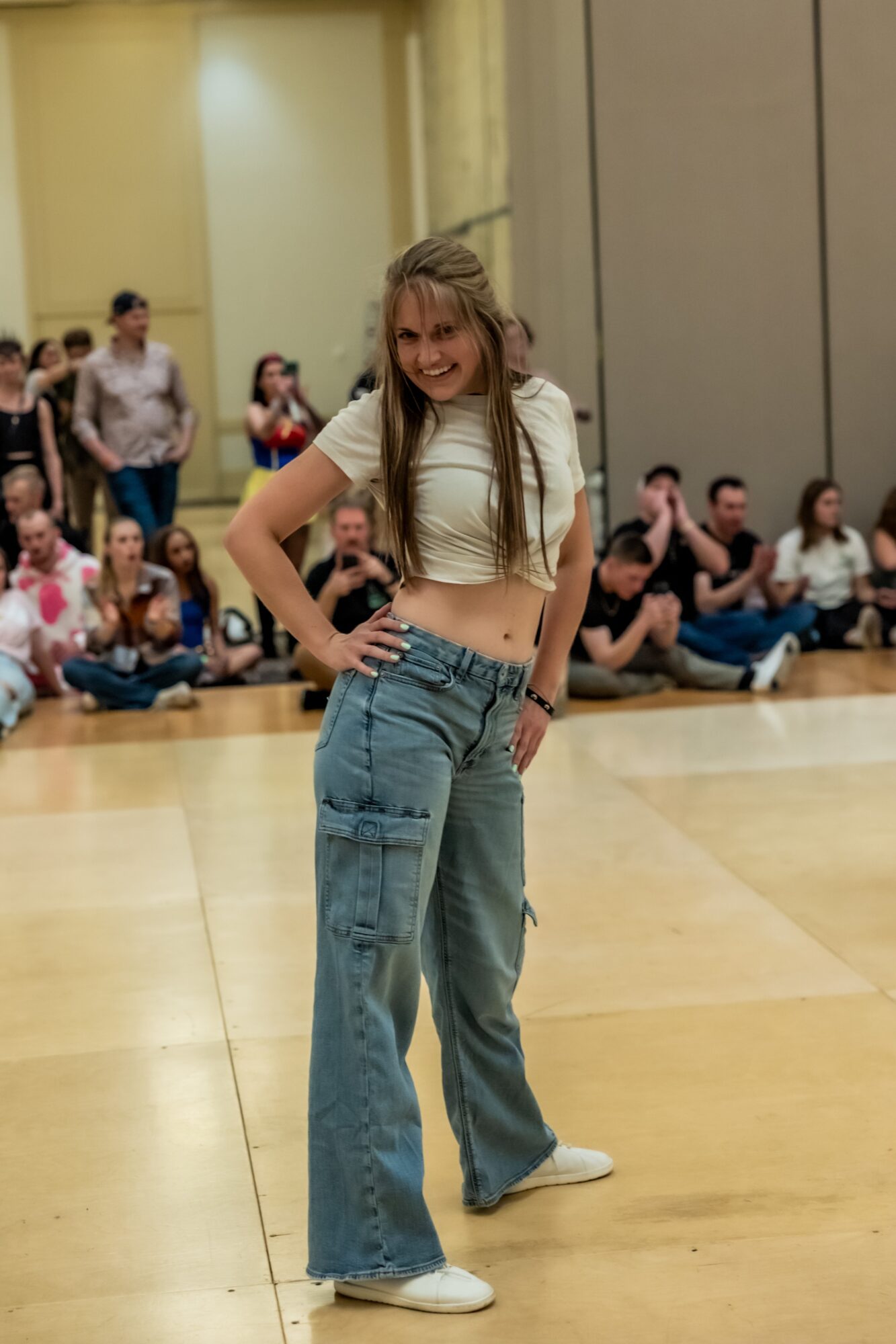 Young woman with long hair in a white crop top and baggy jeans standing on a dance floor, smiling, with people sitting and standing in the background.