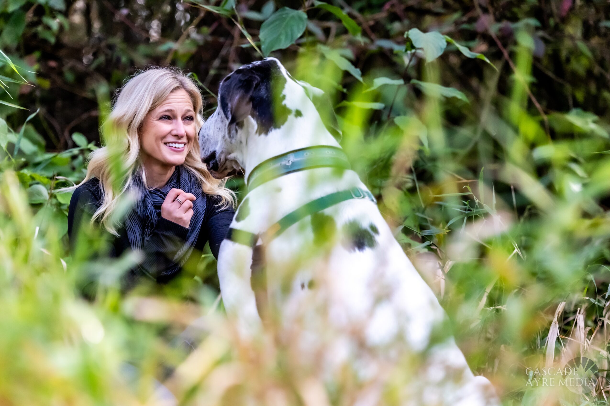 Woman smiling at a large dog in a green outdoor setting, surrounded by foliage.