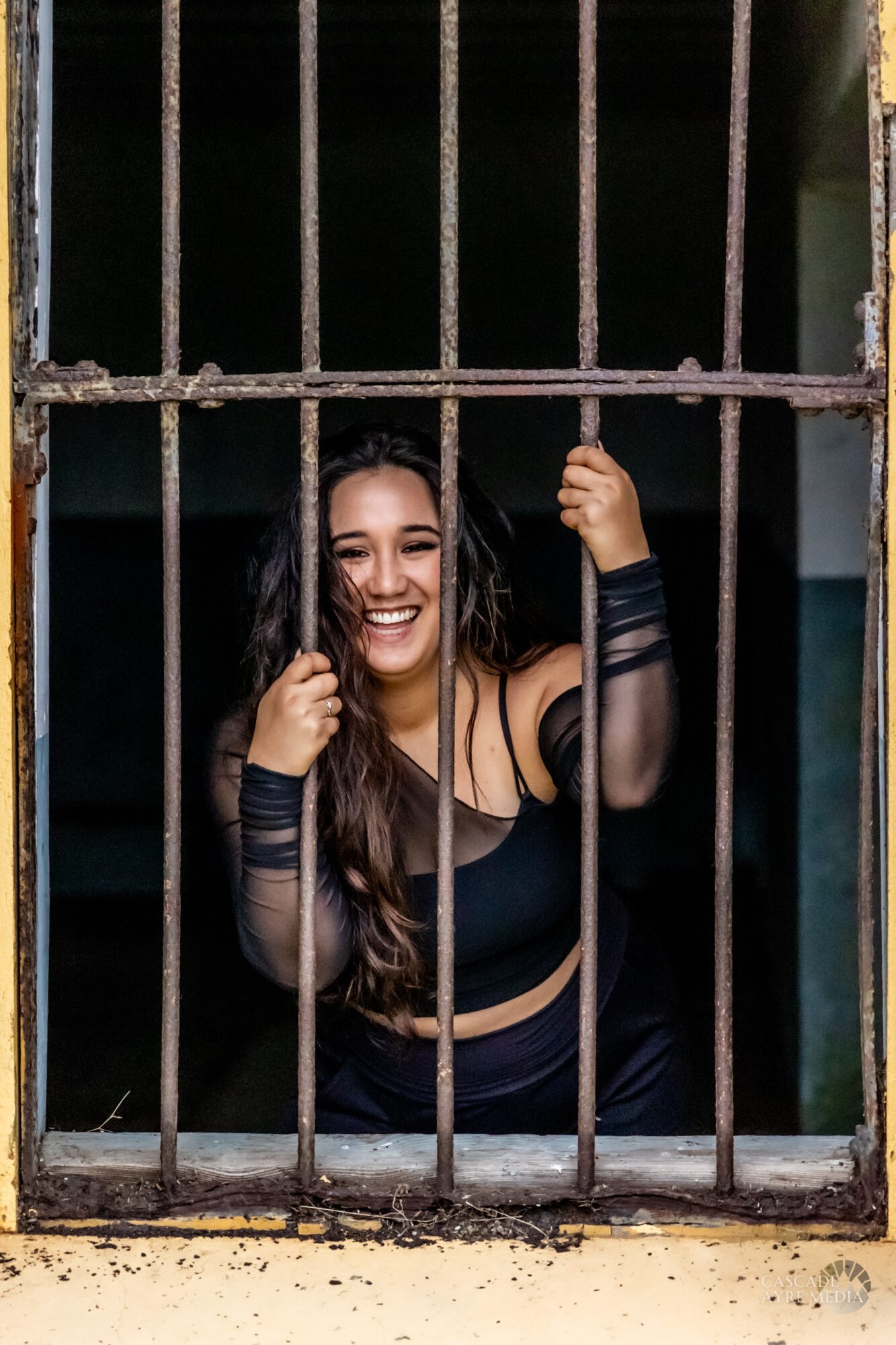 Smiling woman with long hair behind bars, holding one bar with her right hand, in a dark setting.