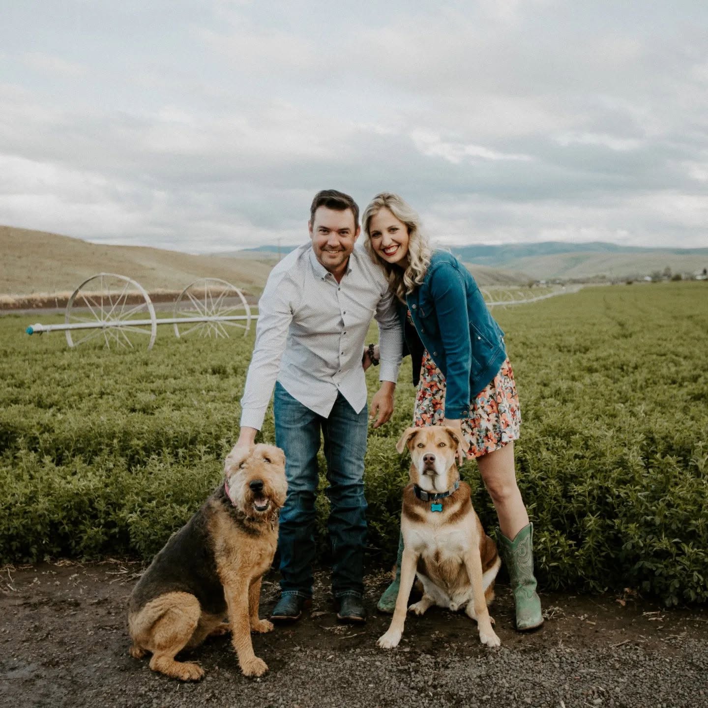 A smiling couple with two dogs outdoors in a green field, cloudy sky above, holding the dogs' leashes.
