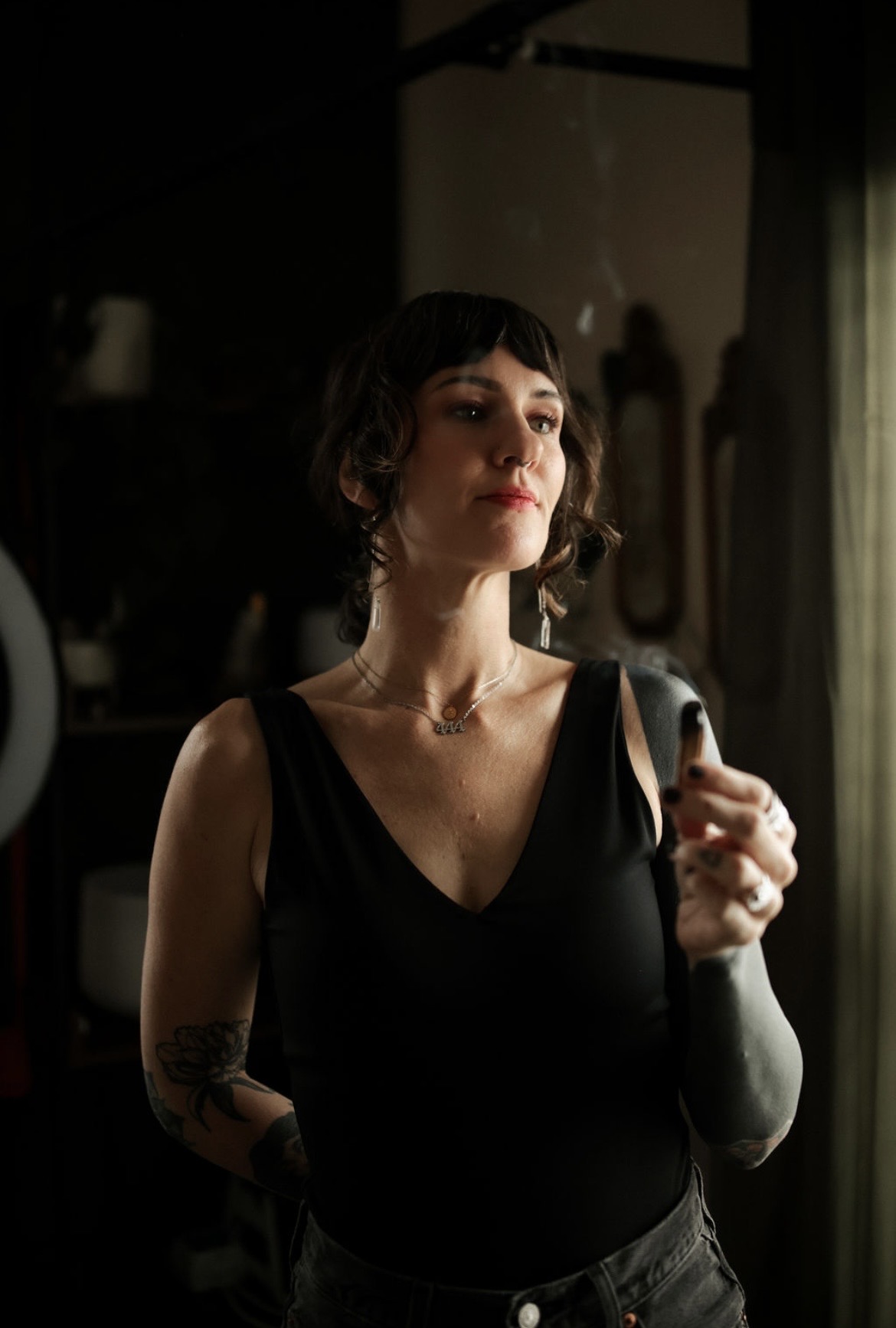 Woman with short dark hair wearing a black sleeveless top, standing near a window with curtains, holding a coat.
