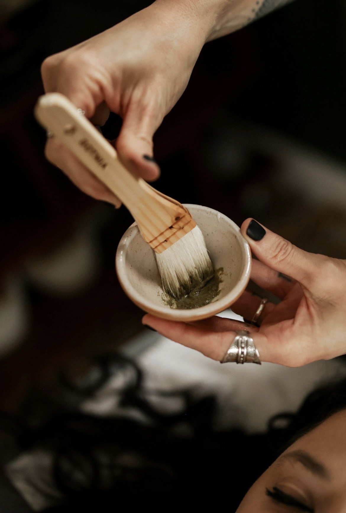 Person holding a small bowl with a brush applying a green substance, with another hand supporting the bowl, rings visible on fingers.