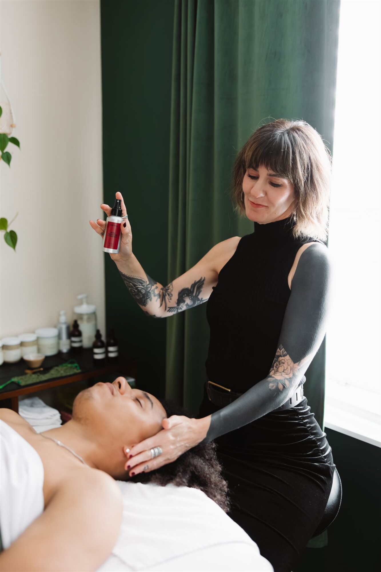 Woman with tattoos holds a syringe near a person lying in bed, in a room with green curtains and natural light.