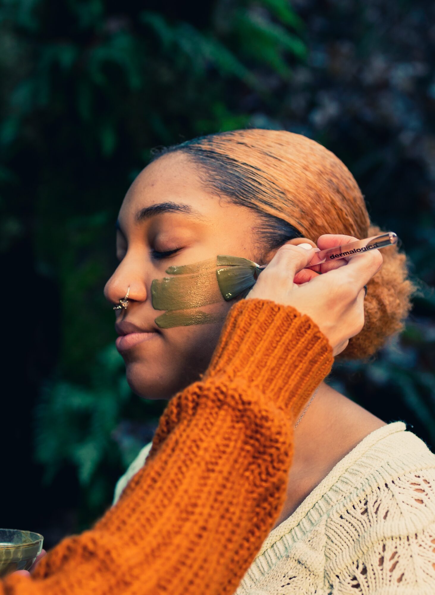 Young woman with closed eyes and braided hair, holding a paintbrush near her face, outdoors with greenery in background.