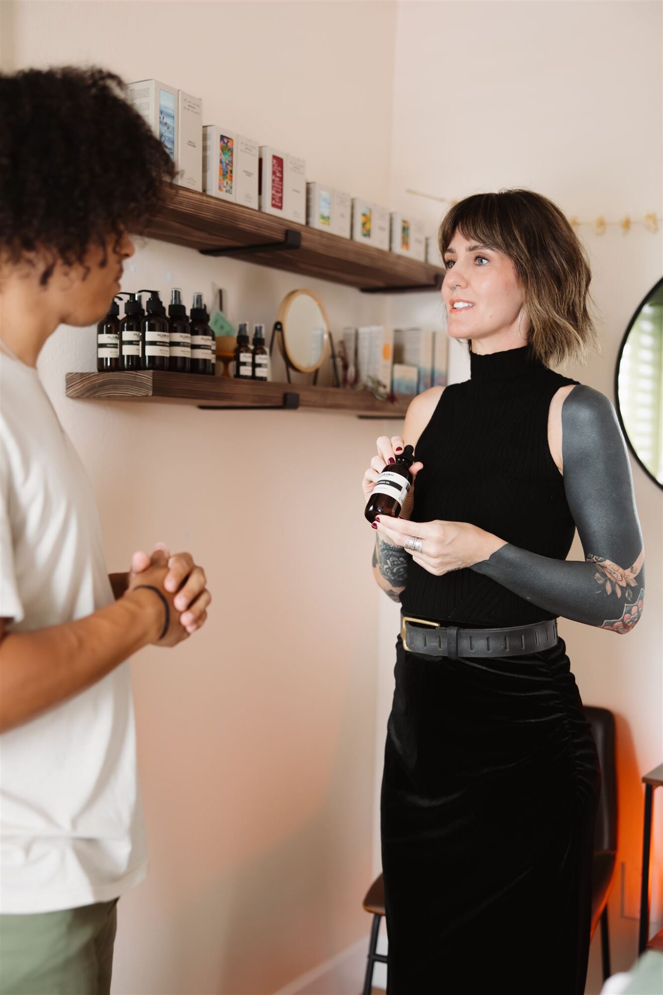 Two women talking indoors, one with curly hair and the other with straight hair and tattoos, holding a small object.
