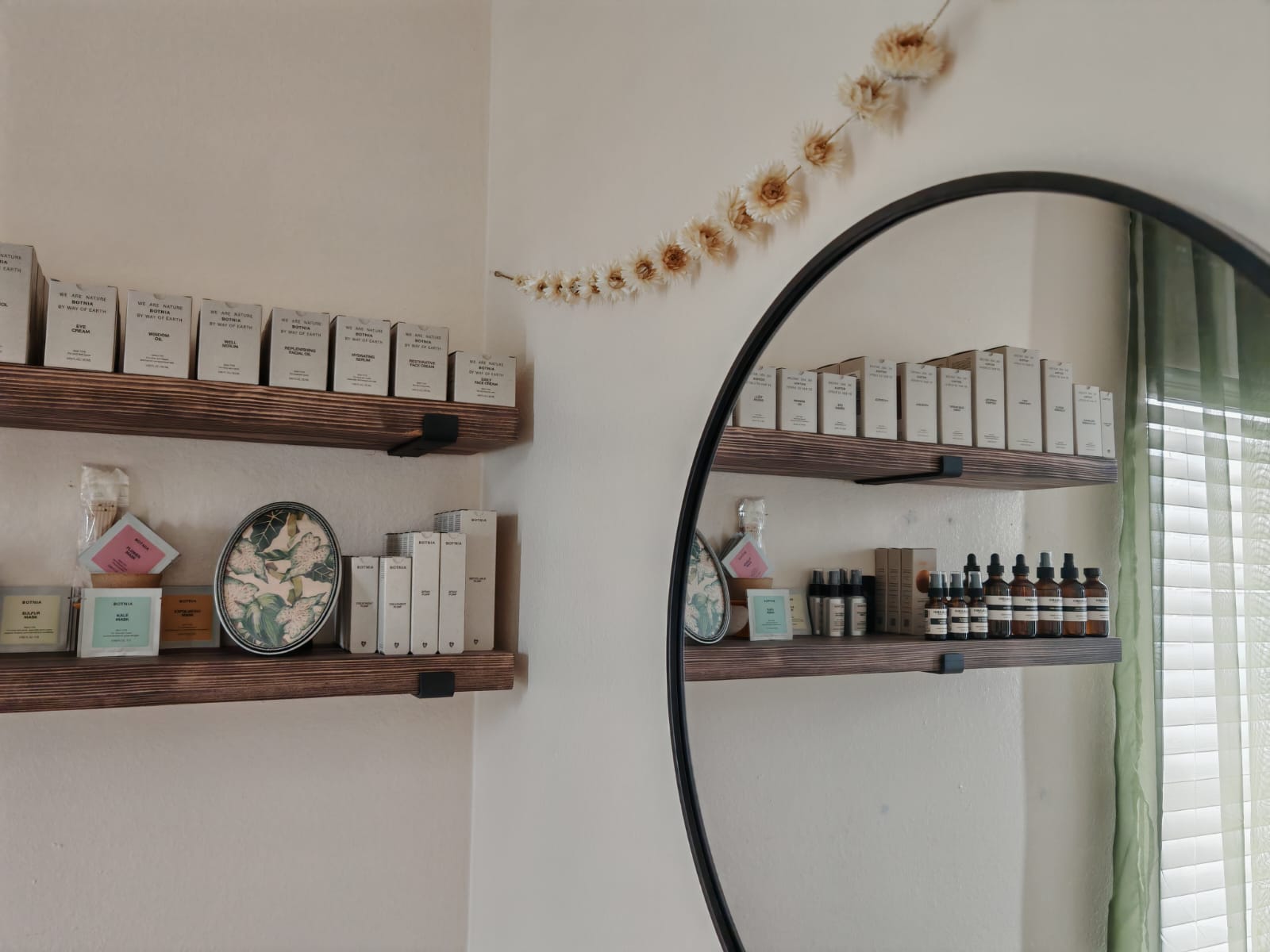 Interior wall with two wooden shelves holding boxes, bottles, and decorative items, reflected in a large round mirror.