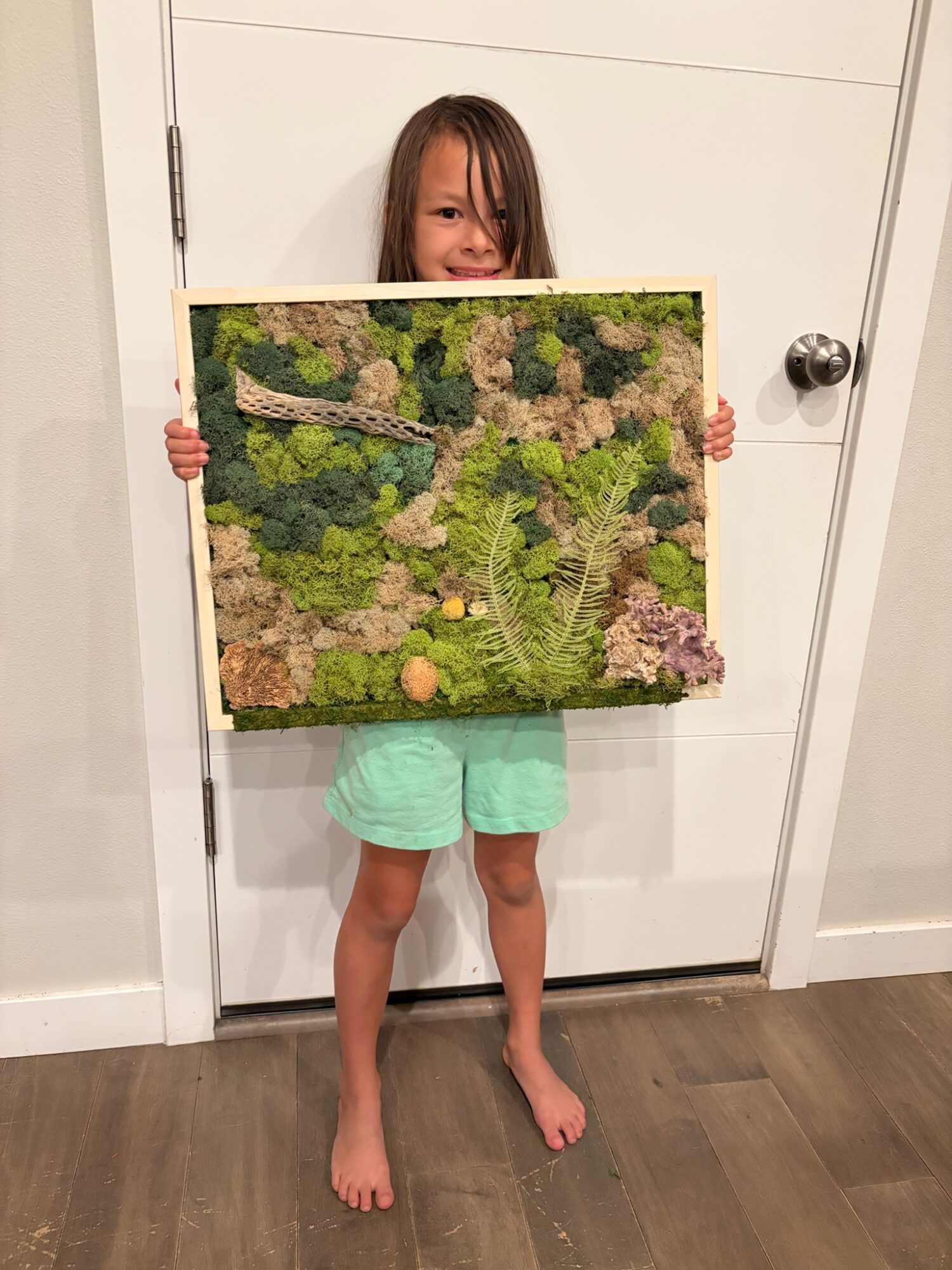 Child holding a framed picture of a forest scene with trees and greenery, standing barefoot in front of a white door.