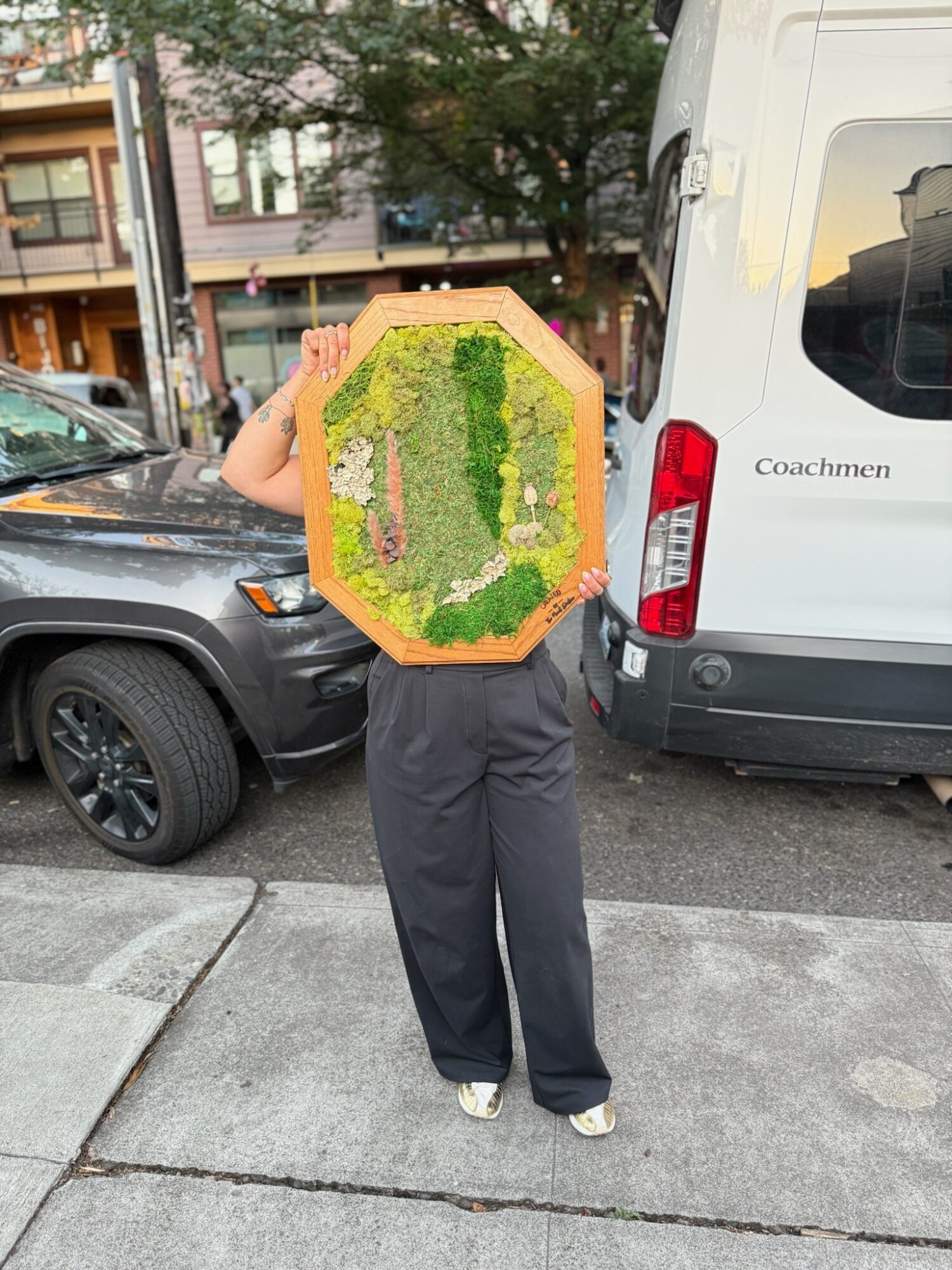 Person holding a large, colorful, hexagonal floral arrangement in front of their face on a city sidewalk.